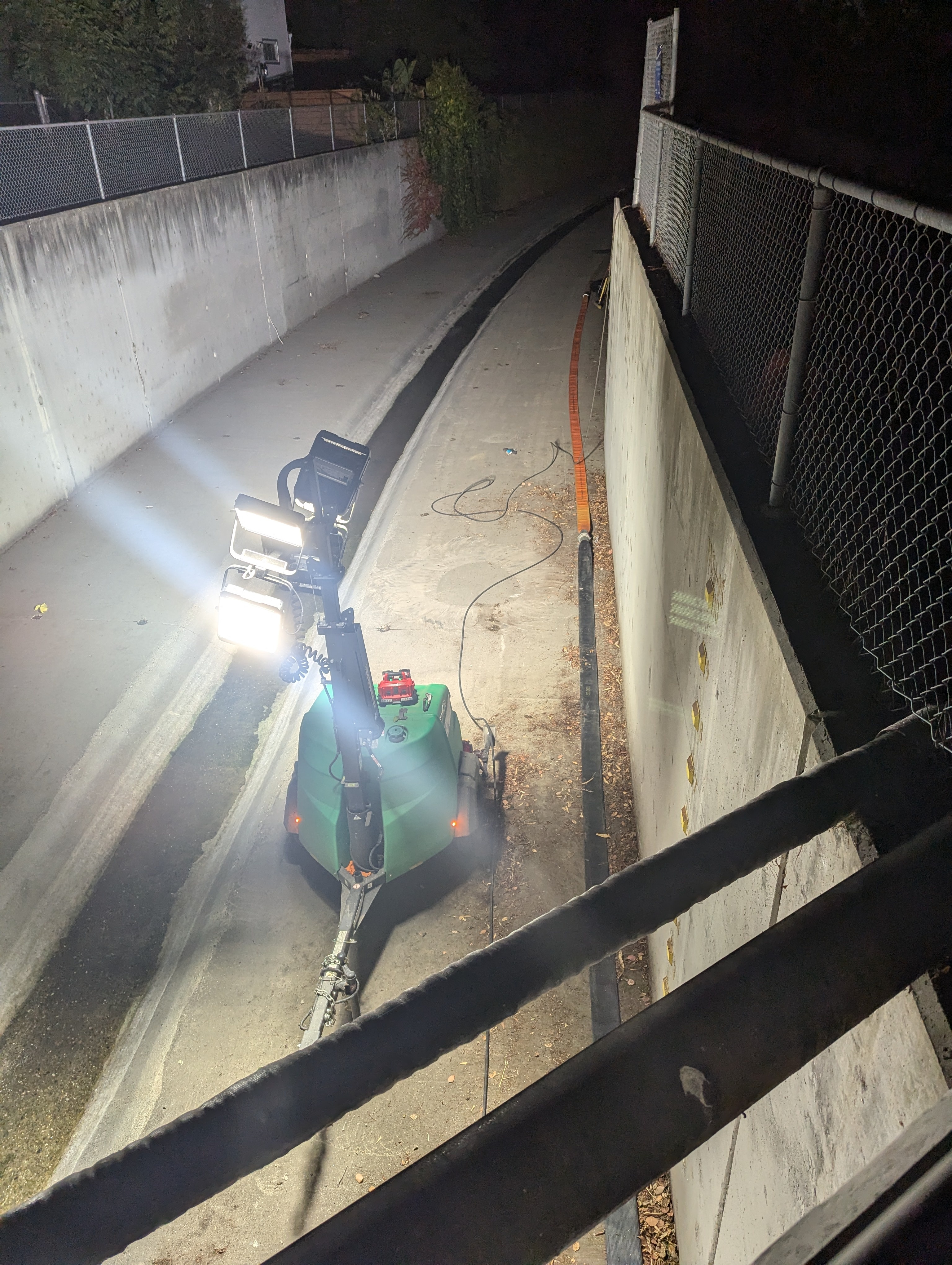 A narrow concrete pathway lit by a bright mobile light machine. It's lined with fences and has cables and an orange hose running along it.