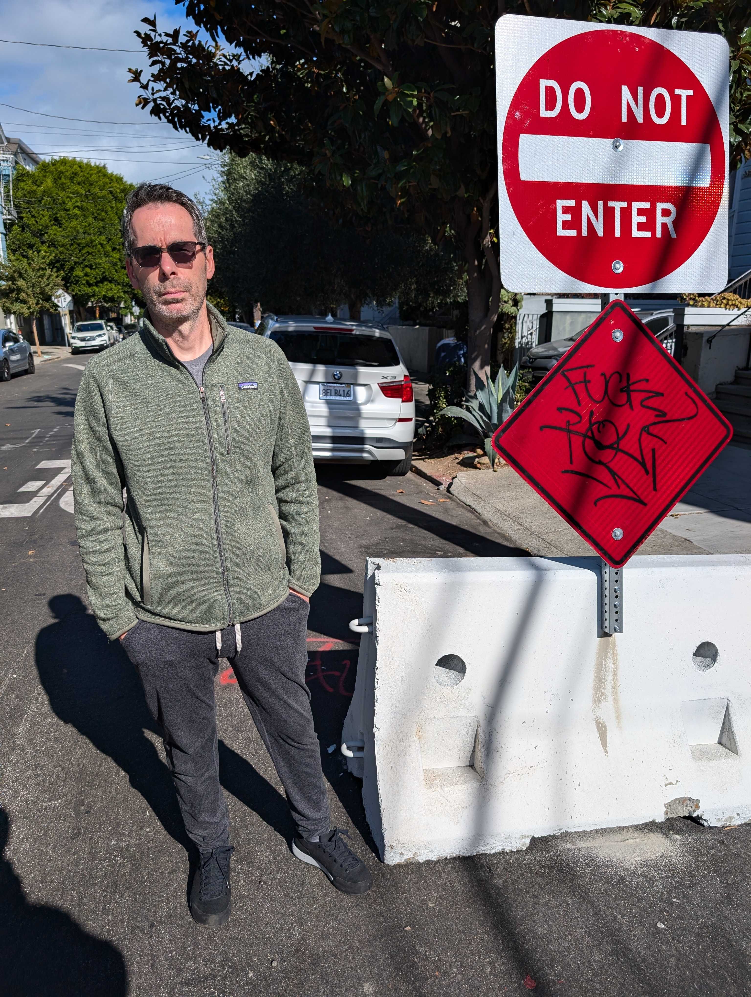 A person in sunglasses stands beside a "Do Not Enter" sign on a sunny street. They wear a green jacket and dark pants, with a car and trees in the background.