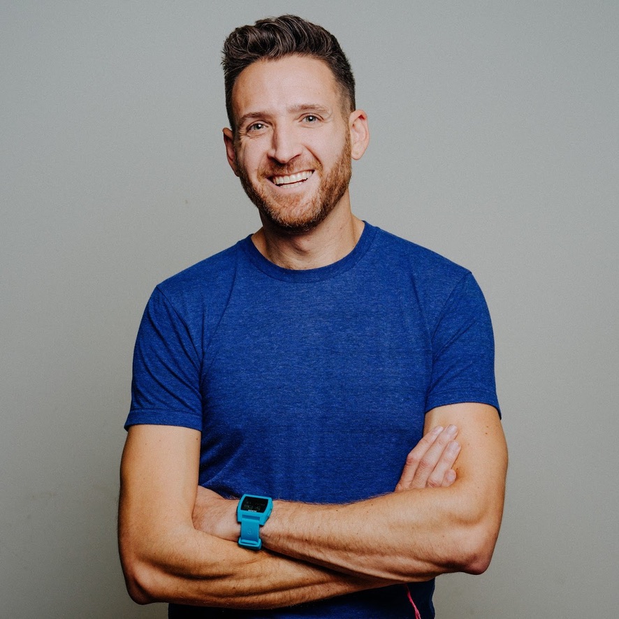 A man smiling with arms crossed, wearing a blue T-shirt and a matching watch, stands against a plain light gray background.