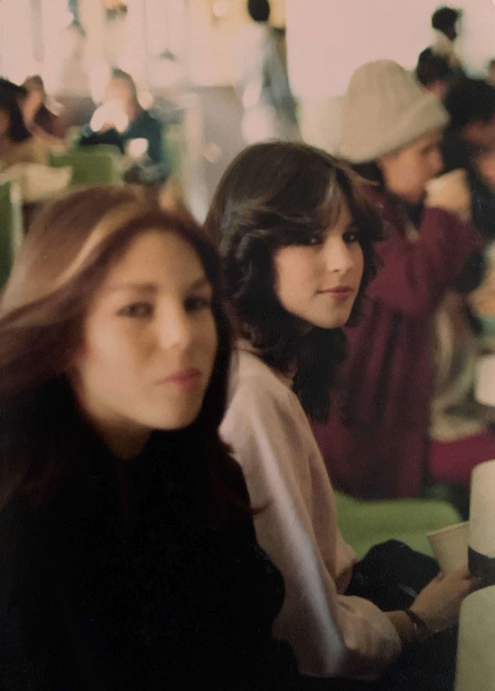 Two young women sit together in a busy indoor setting. One is wearing pink, the other dark clothes, both with long hair. People and tables are blurred in the background.