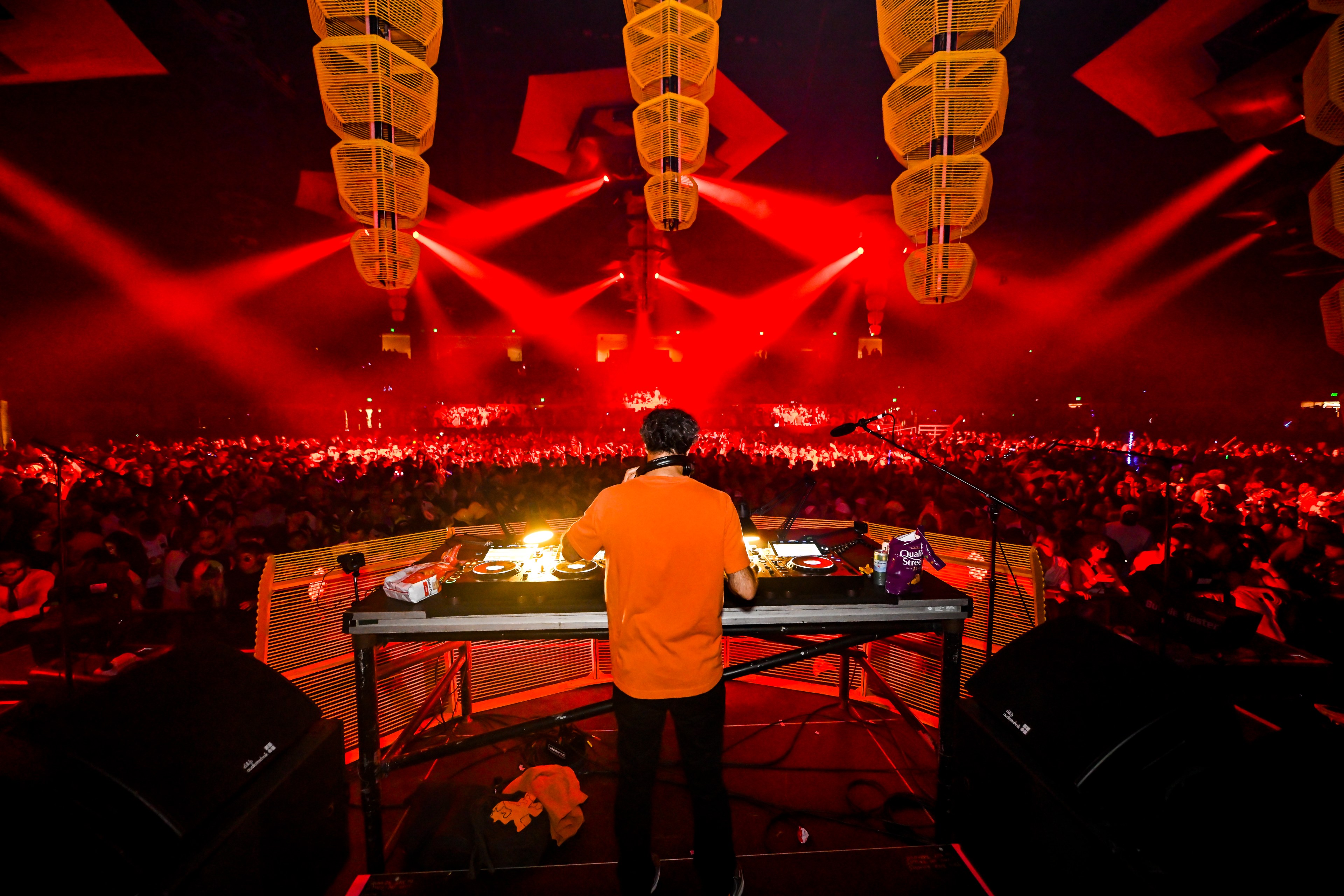 A DJ in an orange shirt plays music on stage under red lighting, facing a large, energetic crowd at a lively concert or festival.