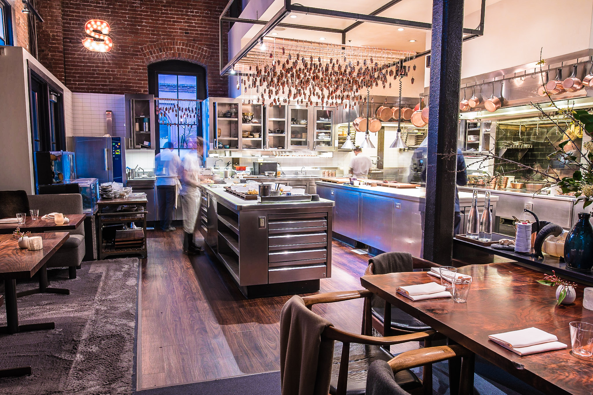 A modern, open kitchen with stainless steel appliances, chefs working, hanging pots, and a cozy dining area with wooden tables and chairs in the foreground.