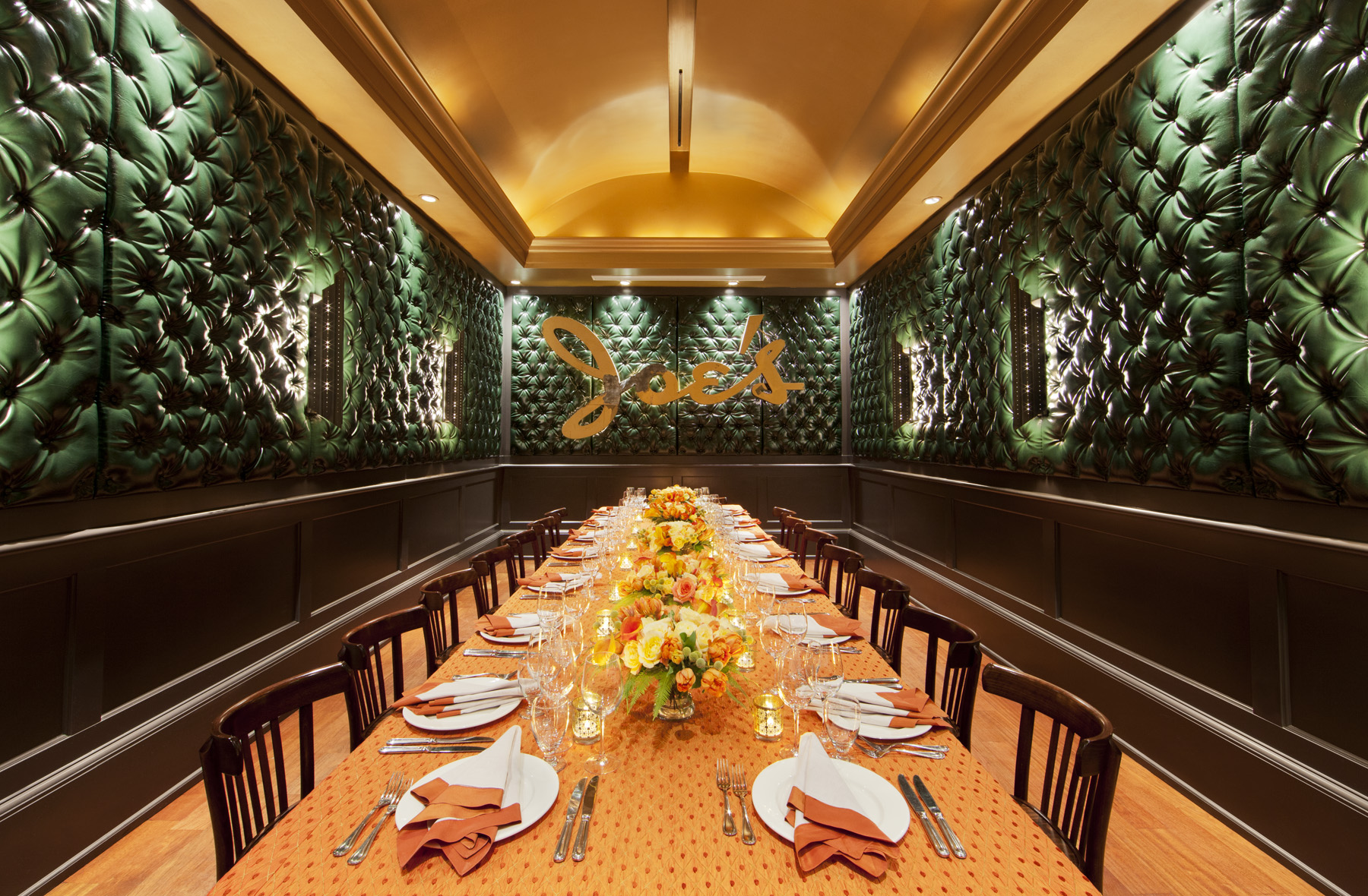 A luxurious dining room features a long table with an orange cloth, set for a meal with elegant dishware and floral centerpieces. Plush green walls add a sophisticated touch.