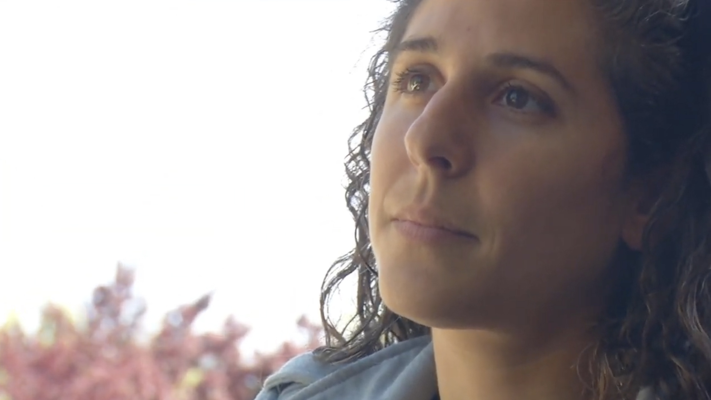 A young woman with curly hair gazes thoughtfully to the side. The background is softly blurred with hints of pink foliage, creating a tranquil atmosphere.