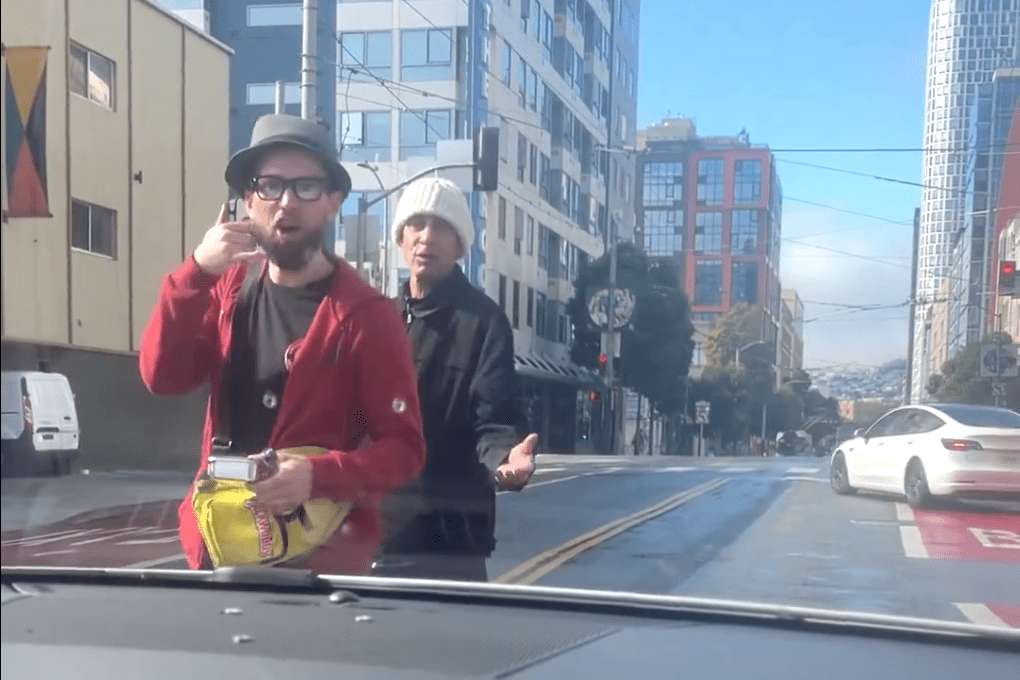 Two men stand in a city street, facing a car. One has a red jacket and hat, holding snacks, making a gesture. The other wears a white beanie. Buildings and cars are visible.