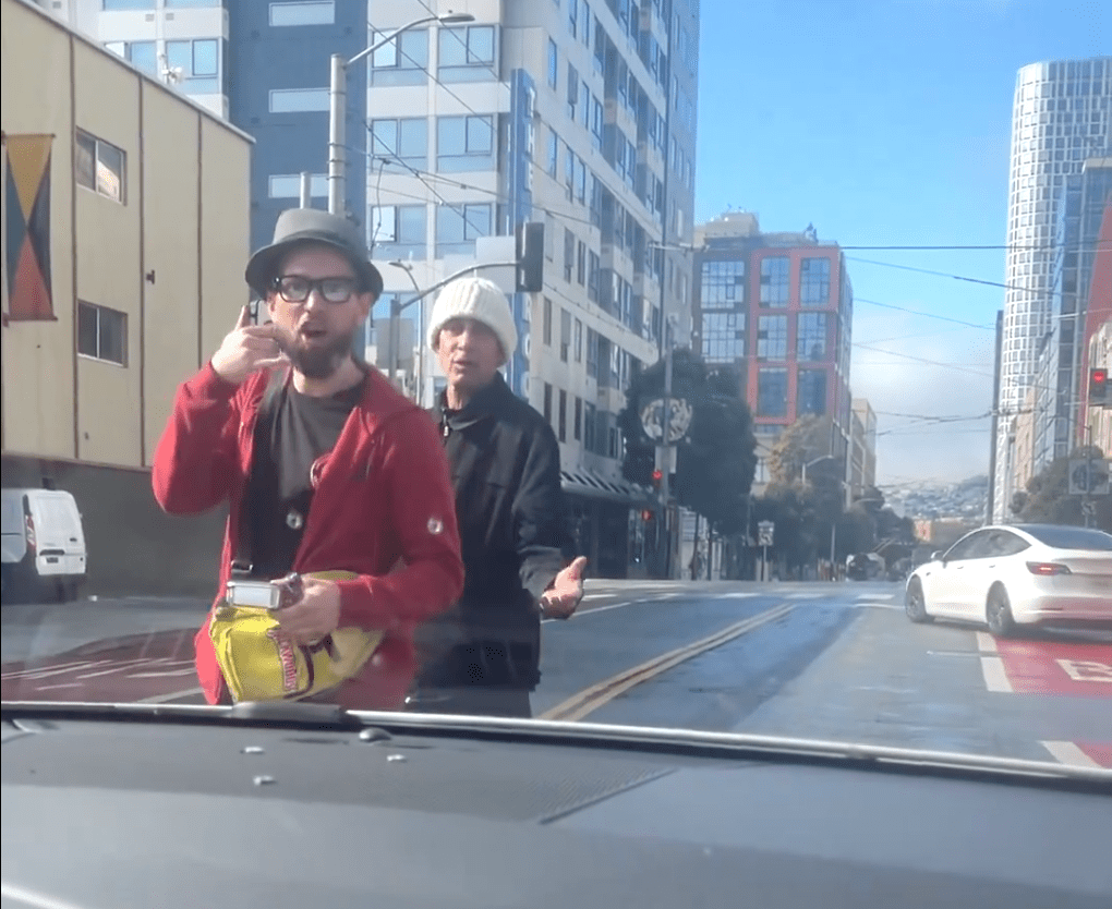 Two men stand in a city street, facing a car. One has a red jacket and hat, holding snacks, making a gesture. The other wears a white beanie. Buildings and cars are visible.