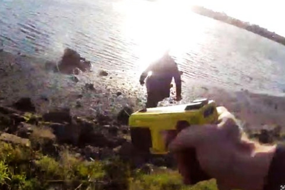 A bodycam view shows a person near a lakefront with a bright glare from the sun reflecting on the water. A yellow Taser is visible in the foreground.