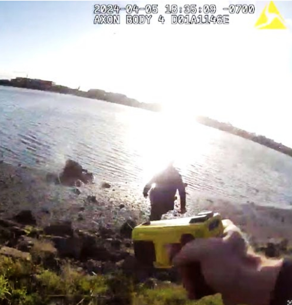 A bodycam view shows a person near a lakefront with a bright glare from the sun reflecting on the water. A yellow Taser is visible in the foreground.