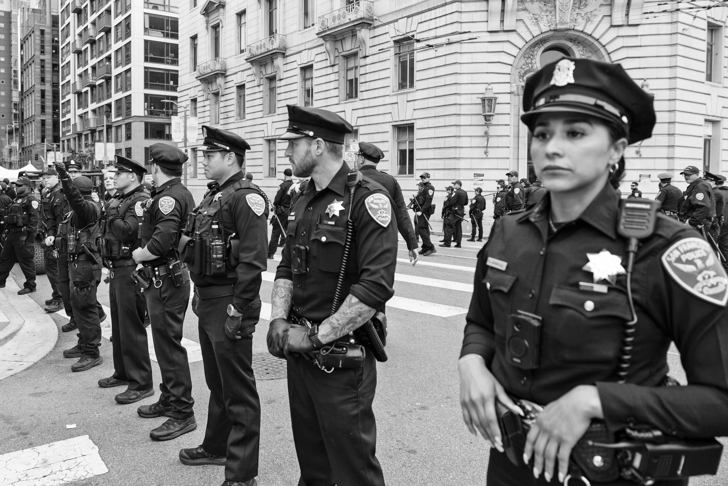 A line of uniformed police officers stands on a city street, each equipped with badges and earpieces, in front of a large building with intricate architecture.