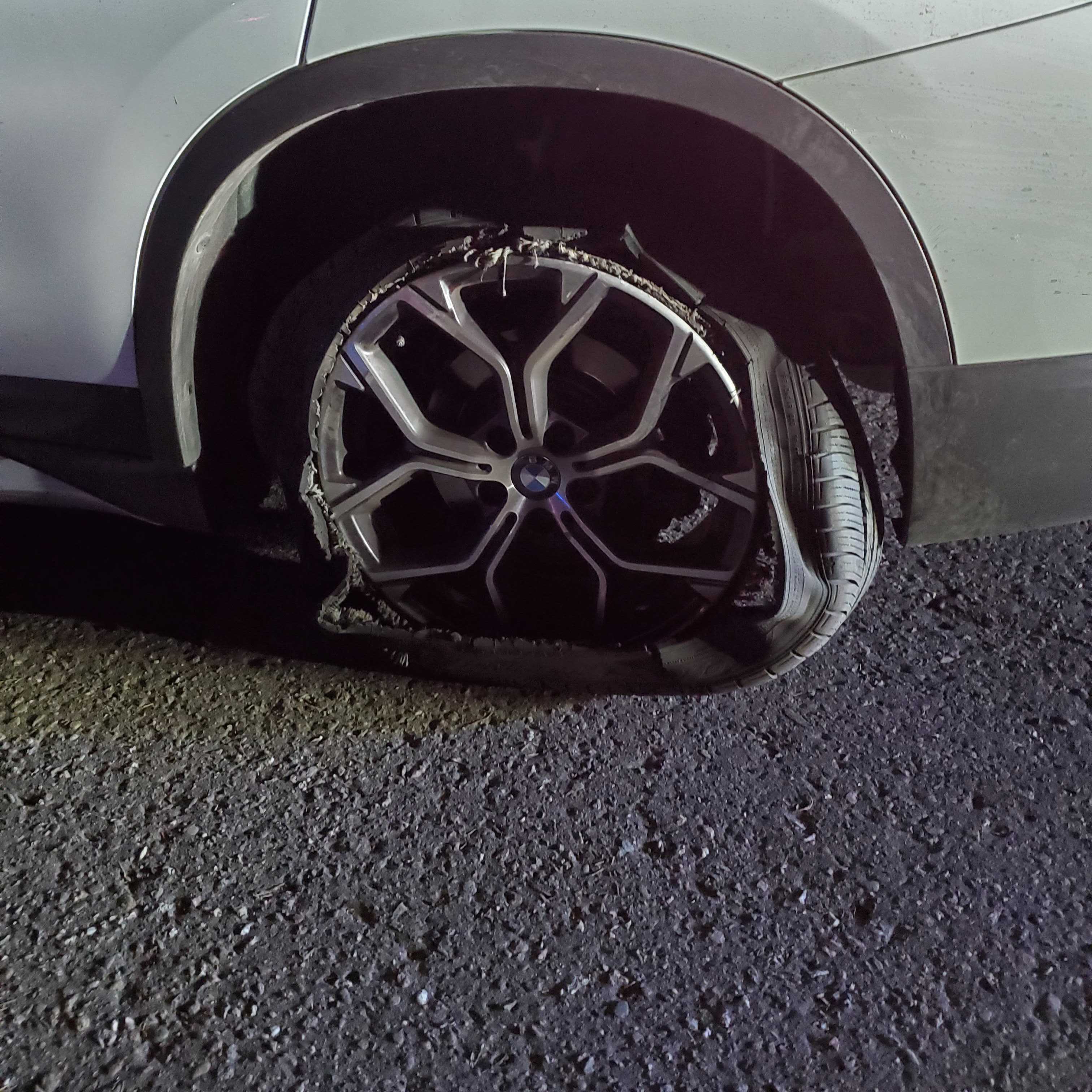 A car tire is severely damaged, with the rubber shredded and hanging off the rim of a BMW wheel. The car is parked on a rough, gravelly surface.