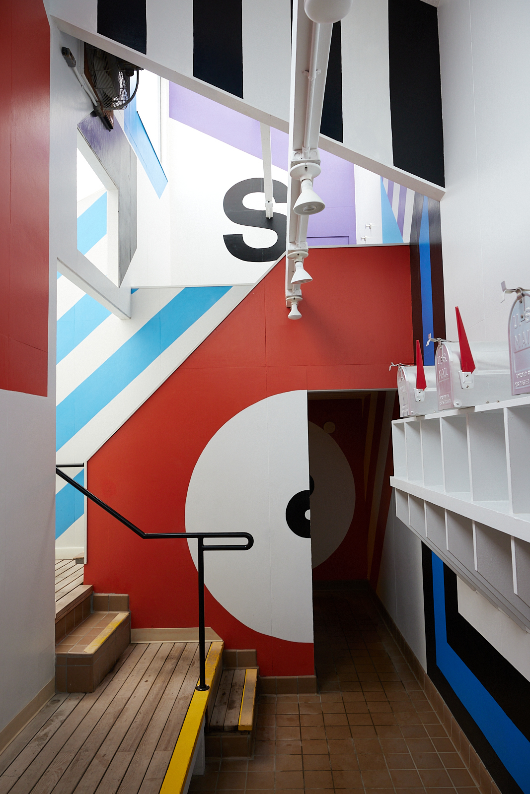 A colorful and geometric stairwell features bold stripes, circles, and letters in red, blue, black, and white. There are white light fixtures above.