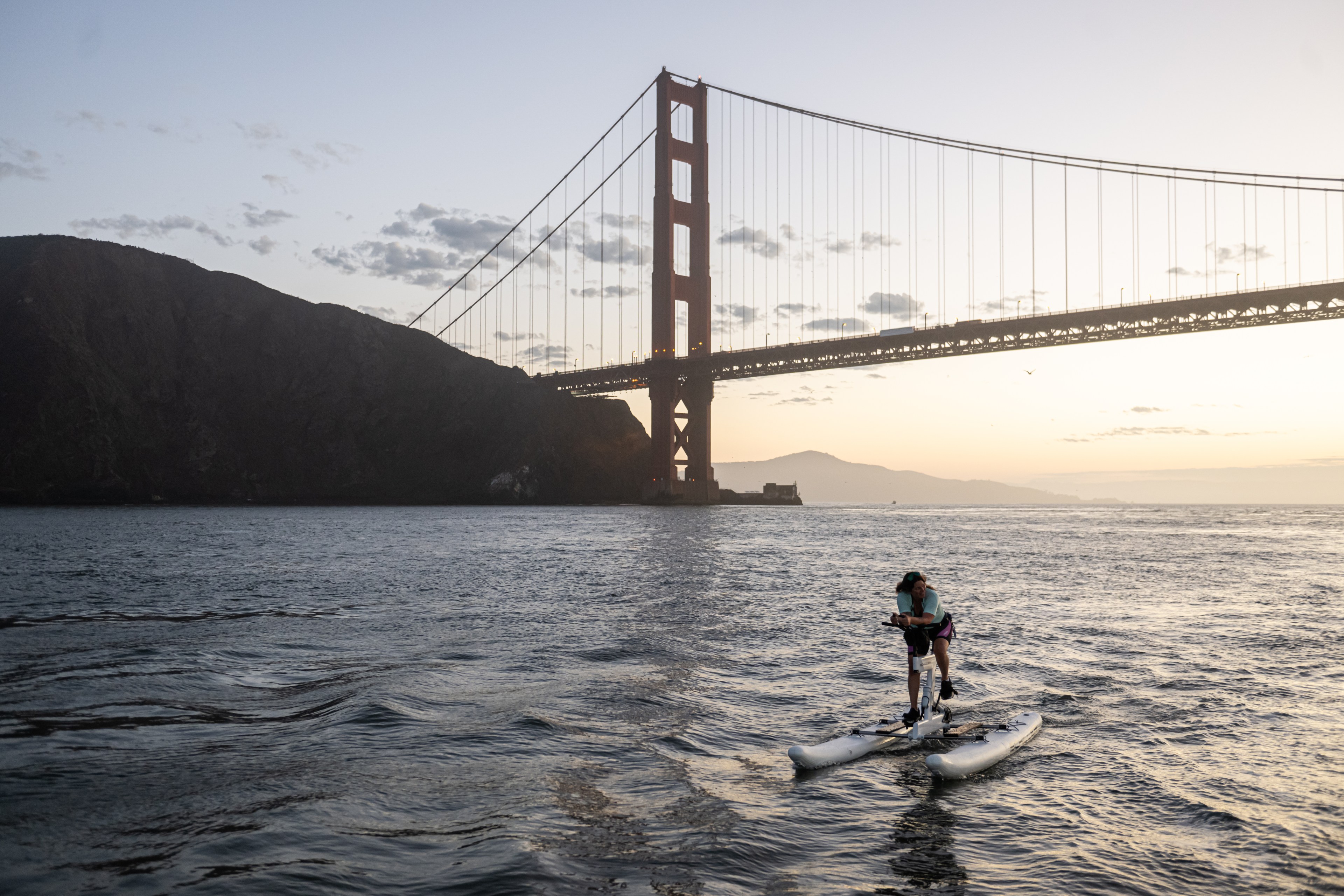 A person rides a water bike under the Golden Gate Bridge at sunset, with dark hills in the background and gentle waves on the water.