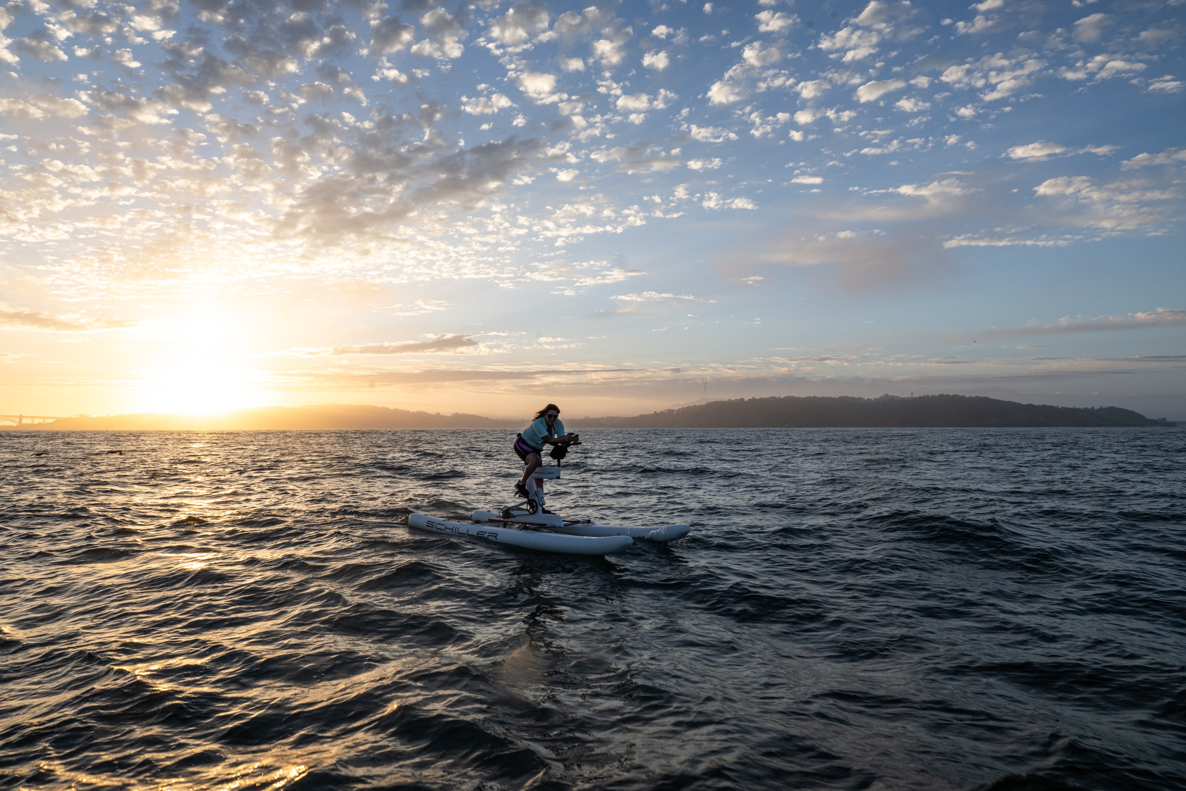 A person rides a water bike on a calm sea during sunset, with a cloudy sky and hills in the background, creating a peaceful and scenic view.