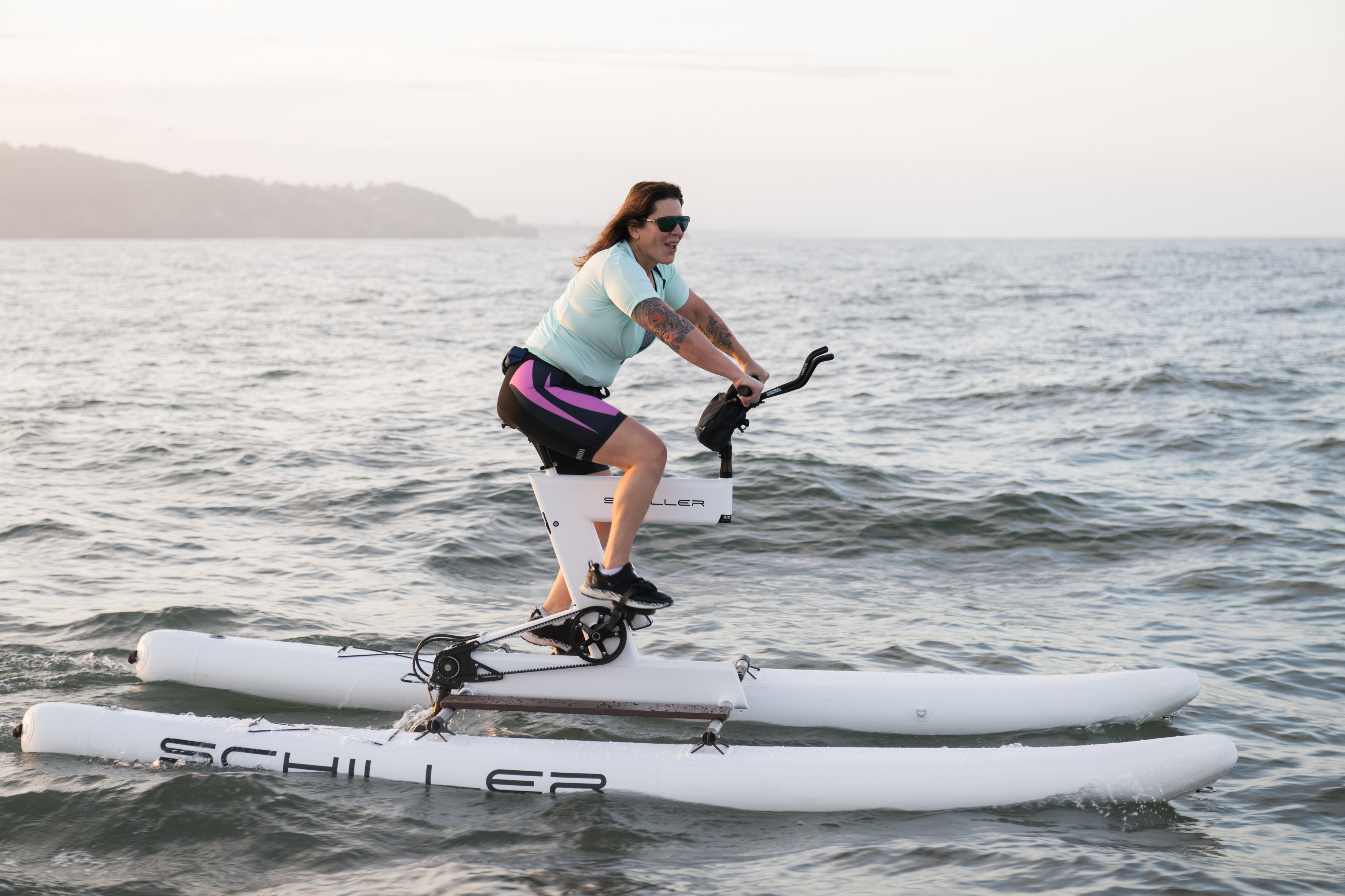 A person is pedaling a water bike on the ocean, wearing a light blue shirt, black and purple shorts, and sunglasses, with land faintly visible in the background.