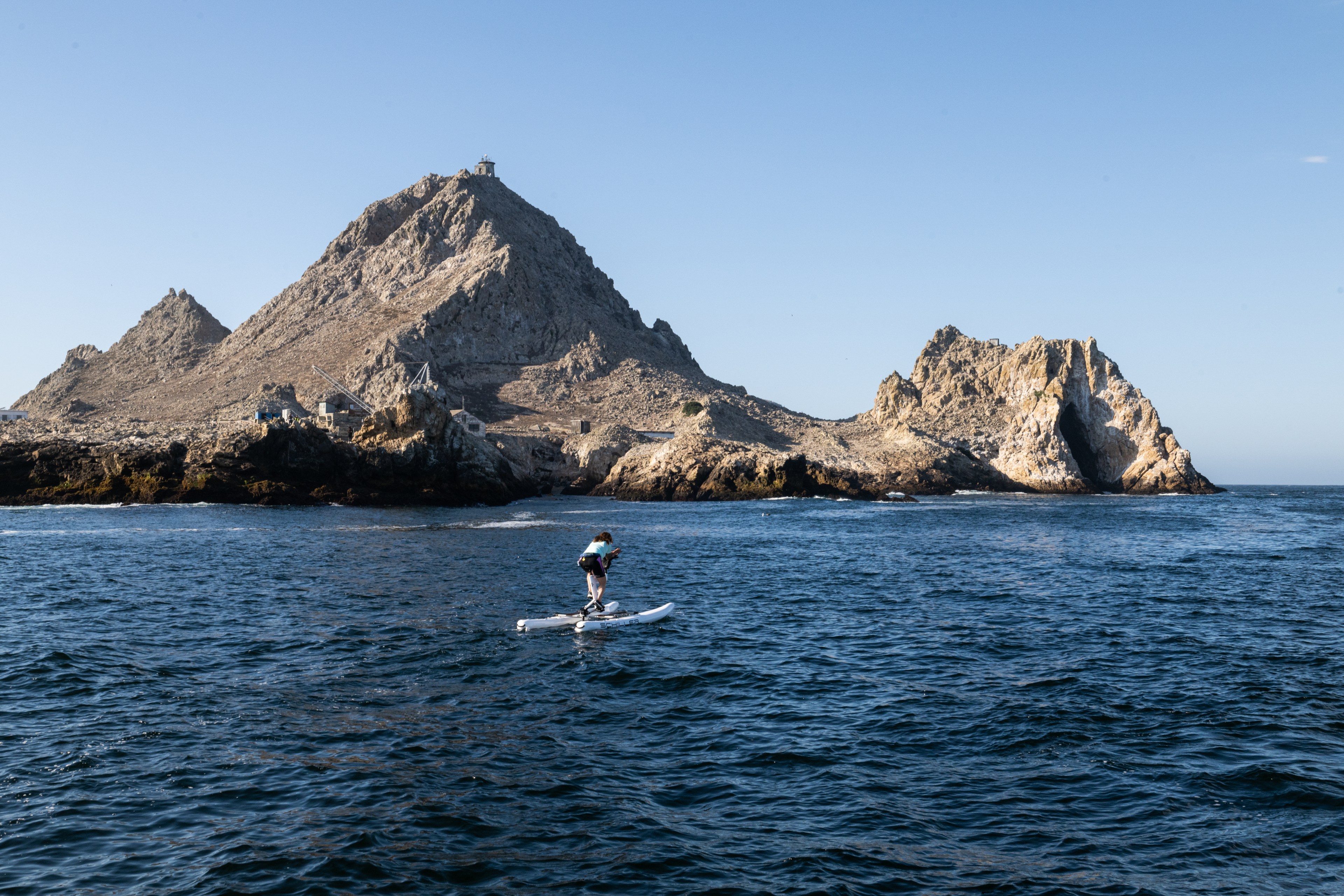 A person rides a pedal-powered watercraft on the blue sea, with rocky islands in the background under a clear blue sky.