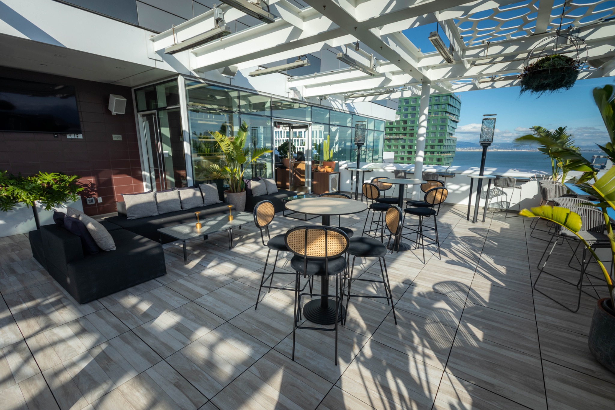 A rooftop terrace features modern seating, tables, and greenery. It overlooks the water with a view of buildings. The area is sunny with partial shade.