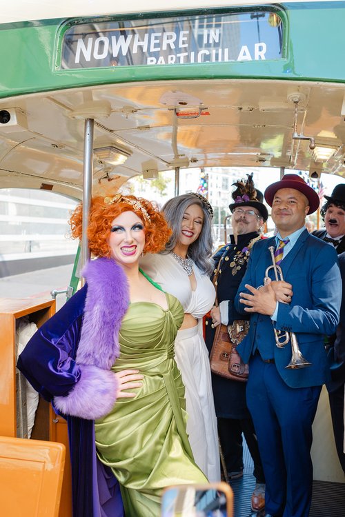 People in vibrant costumes and makeup are on a trolley labeled &quot;NOWHERE IN PARTICULAR.&quot; They are smiling and one person holds a trumpet.