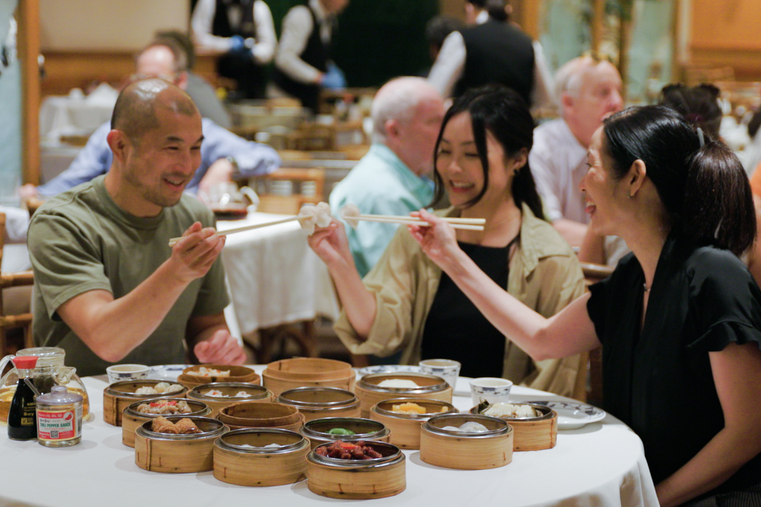 Three people are joyfully sharing dim sum at a restaurant table filled with various bamboo steamers and sauce bottles. They are using chopsticks to pick up food.