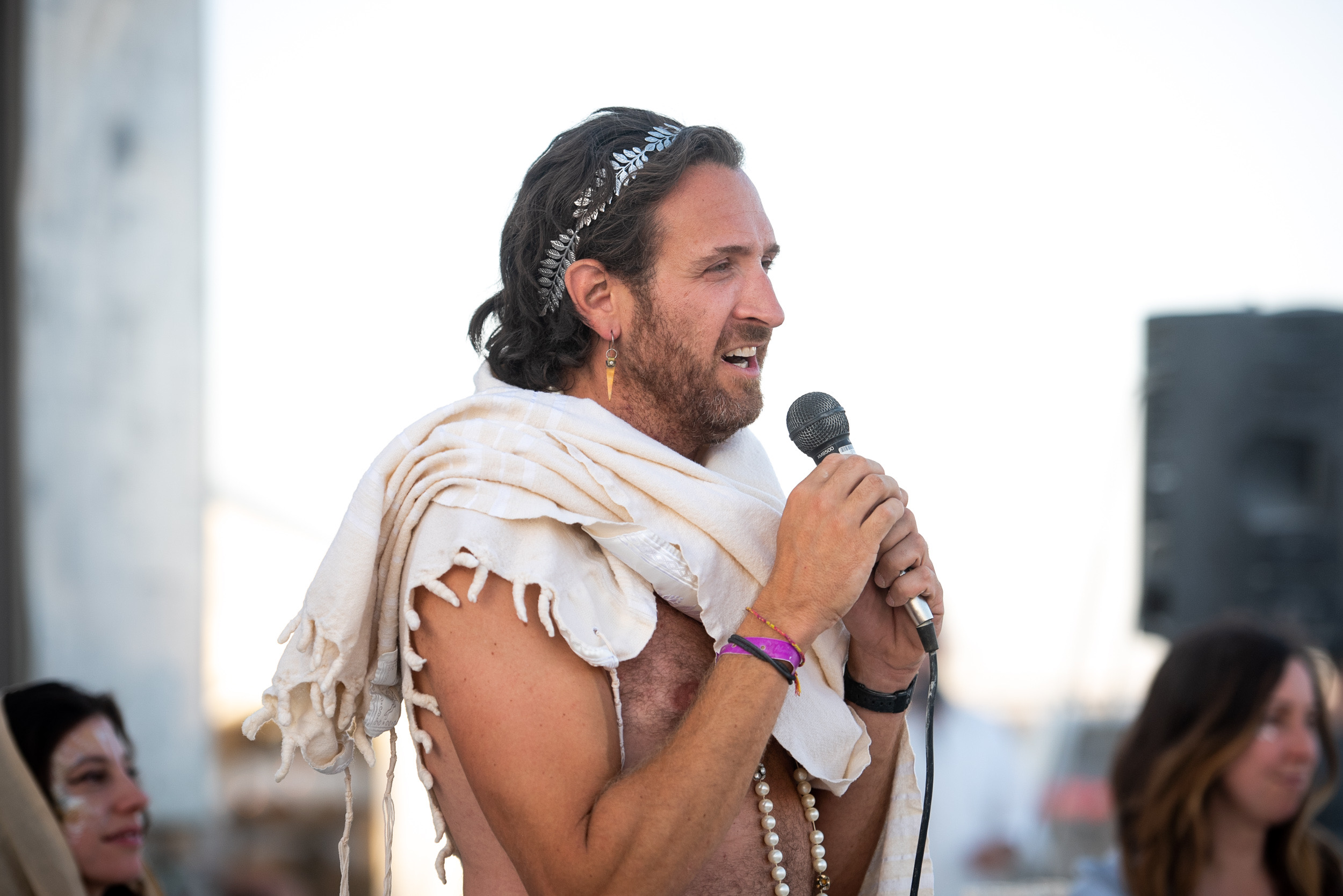 A man holds a microphone while speaking. He wears a headband, light shawl, beaded necklace, and bracelets. There's a blurred figure in the background.