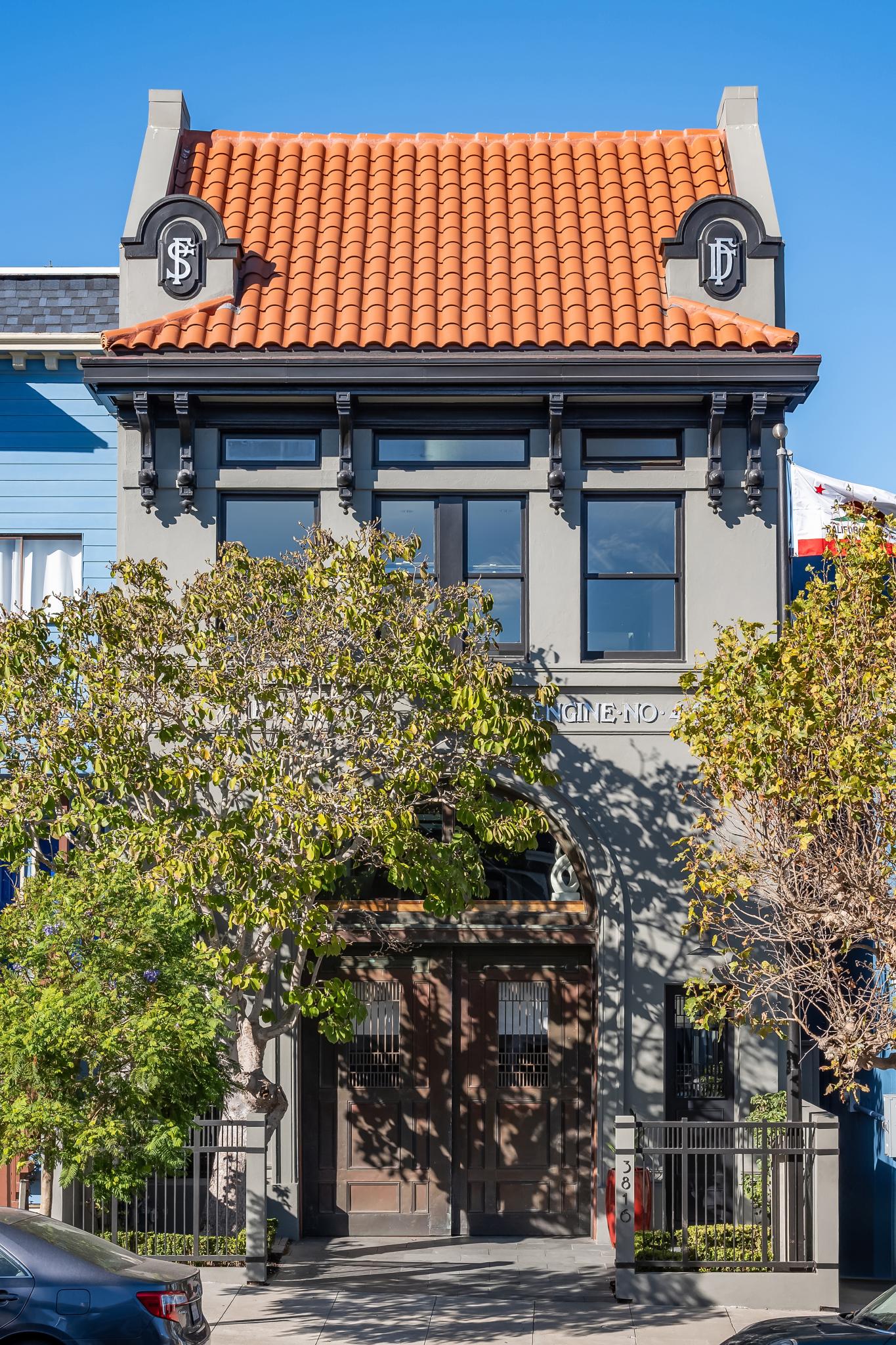 The image shows a two-story building with a red tiled roof and large black-framed windows. Trees partially obscure the wooden entrance. A California flag is visible.