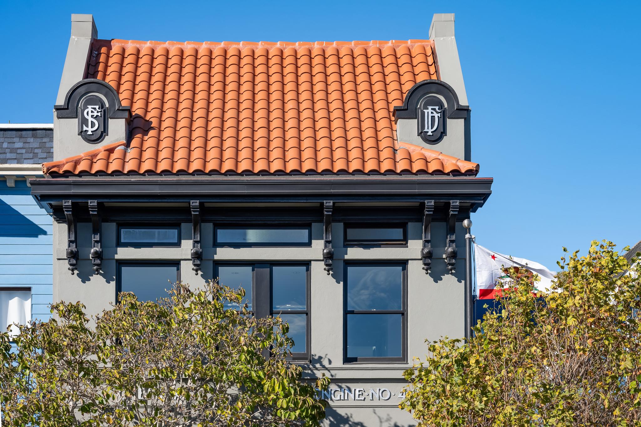 The image shows a building with a red-tiled roof, dark window frames, and ornate design details. Trees partially obscure the lower part, and a flag waves nearby.