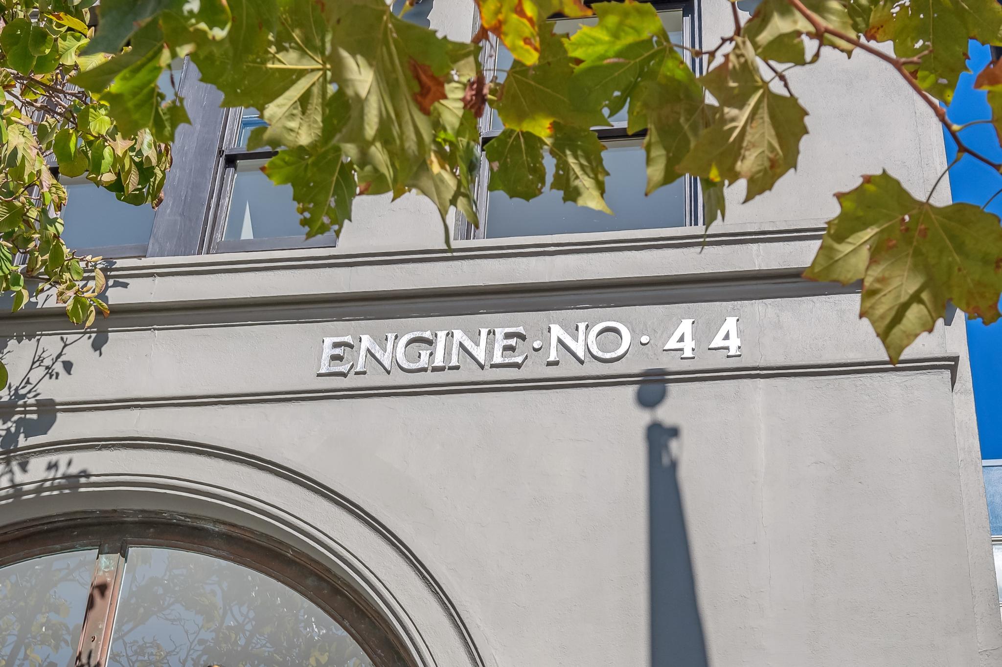 The image shows a gray building facade with &quot;ENGINE NO. 44&quot; in bold white letters. Green leaves partly frame the top, and sunny blue sky is visible.