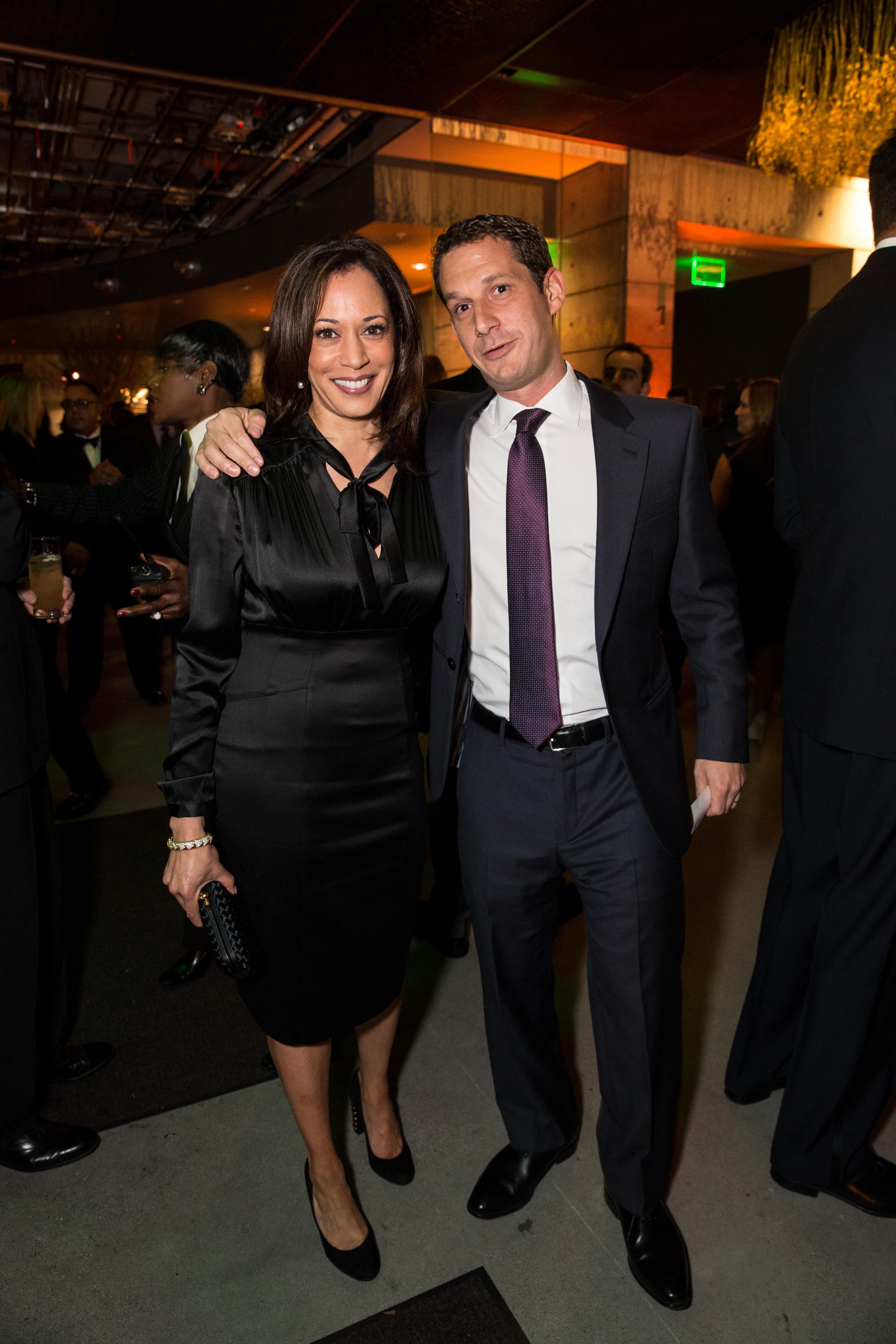 A woman in a black dress and a man in a suit with a tie smile at a social event, surrounded by other formally dressed attendees.