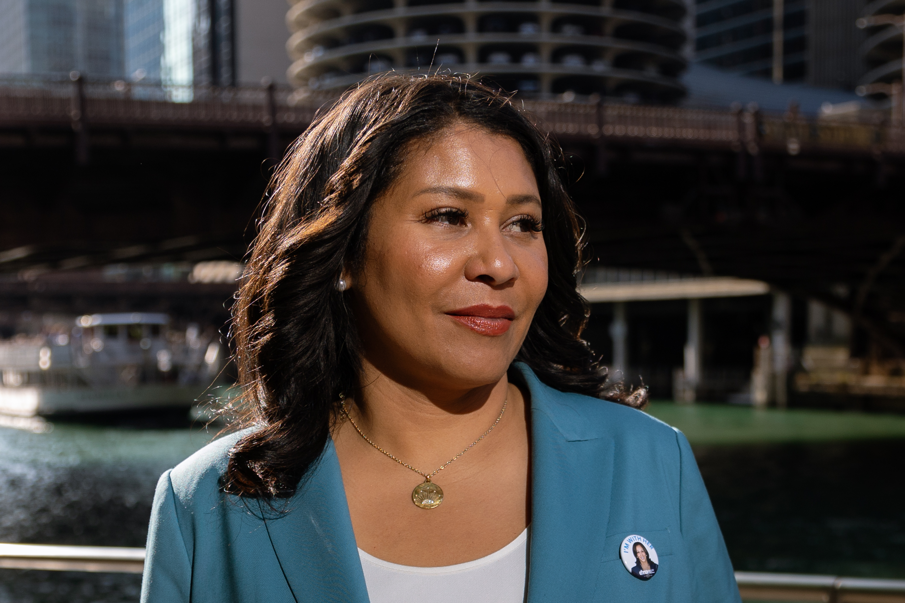 A woman in a blue blazer stands near a river with city buildings in the background. She has a circular pin on her lapel and appears thoughtful.