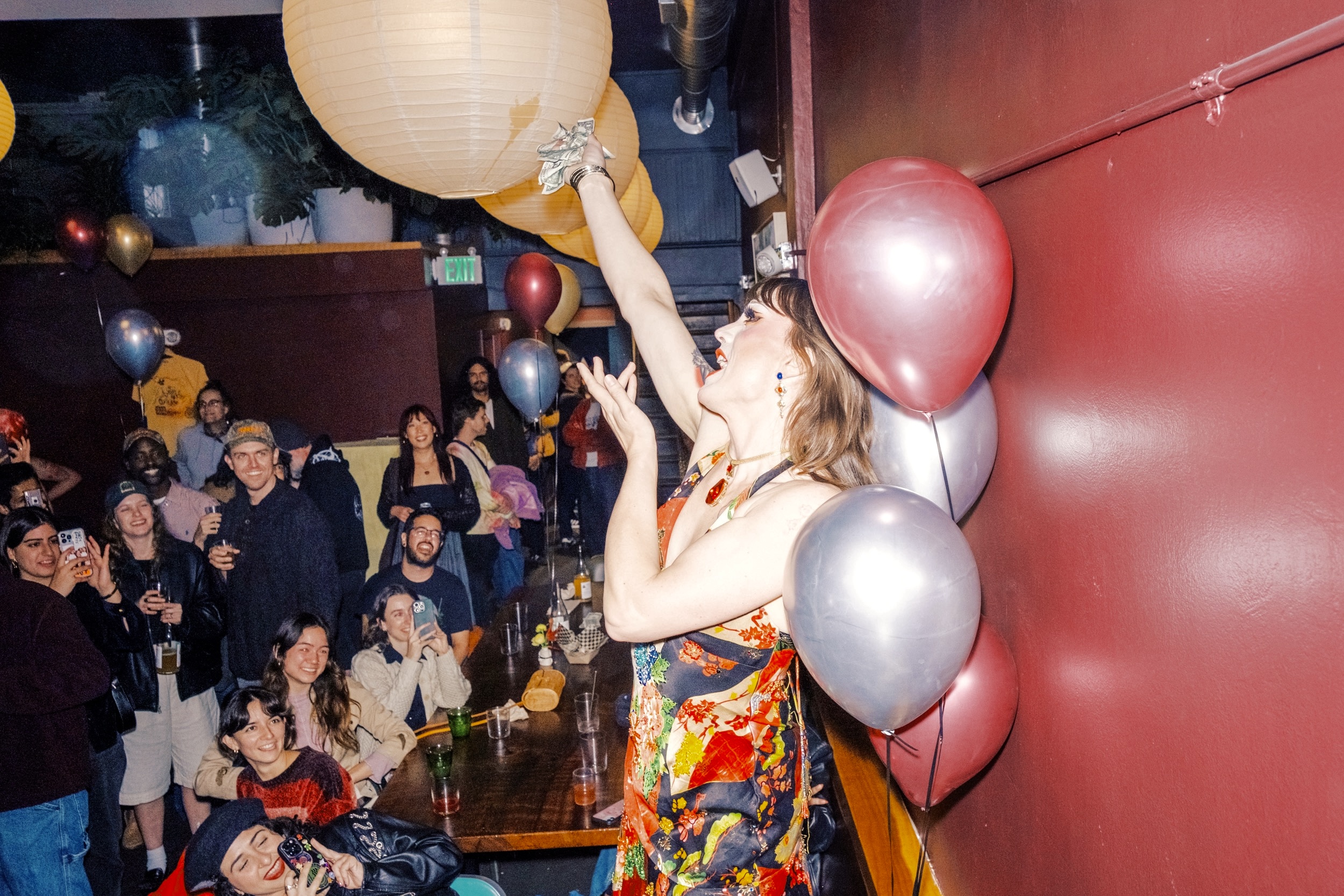 A lively party scene shows a person in a colorful dress reaching for a hanging lantern, surrounded by smiling guests and balloons in a warmly lit room.