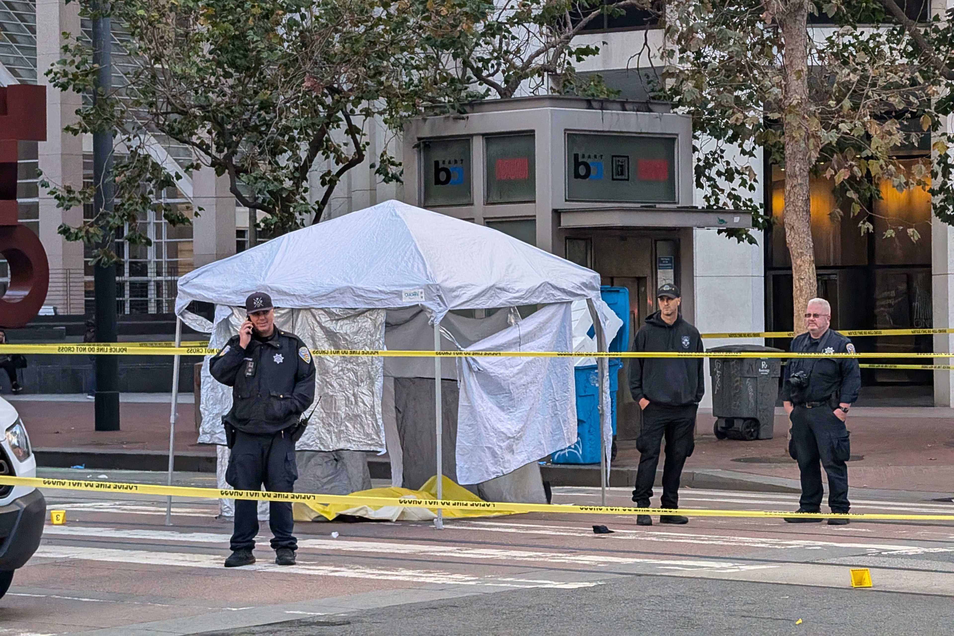 A street scene shows three police officers standing near a white tent, with yellow caution tape cordoning off the area. There are trees and a building in the background.