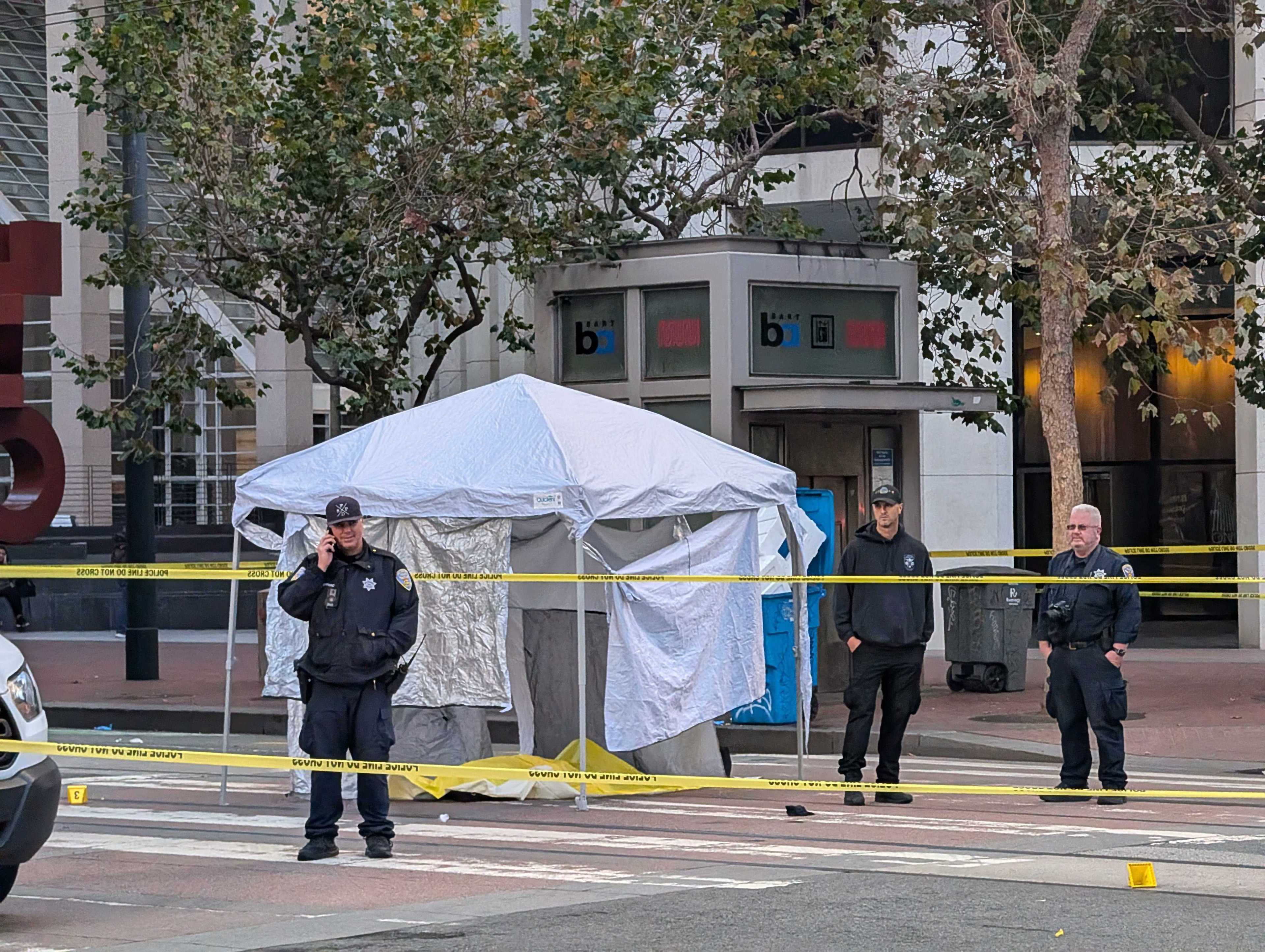 A street scene shows three police officers standing near a white tent, with yellow caution tape cordoning off the area. There are trees and a building in the background.