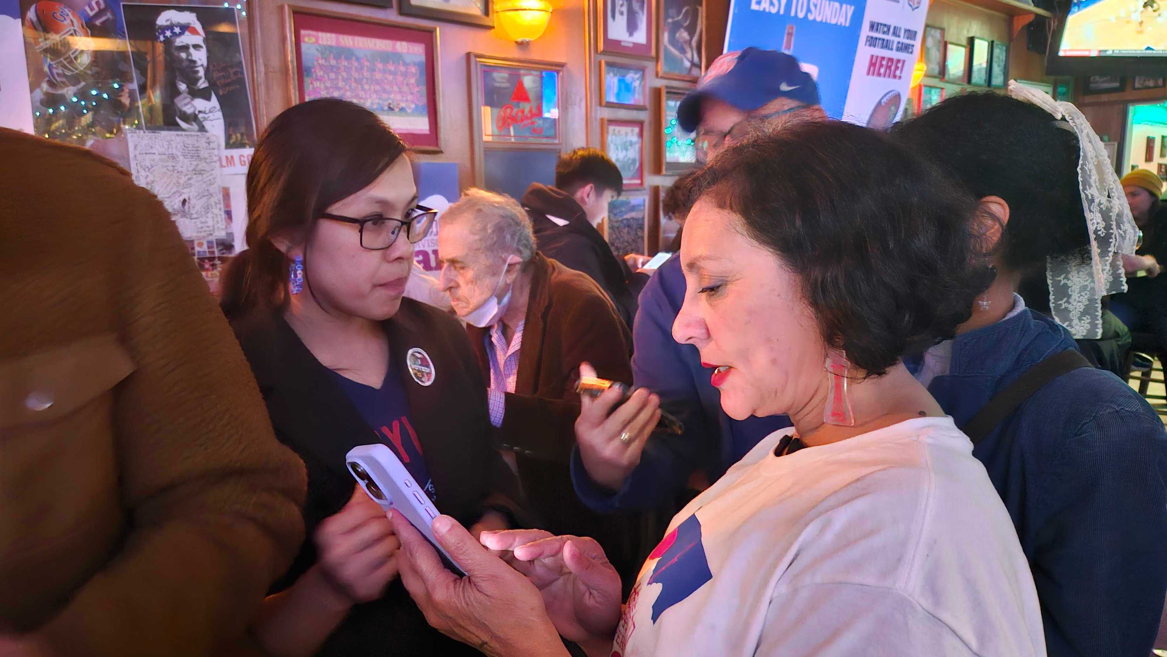 Two women focus on a smartphone in a lively bar, surrounded by others and vibrant wall art, creating an engaging atmosphere.