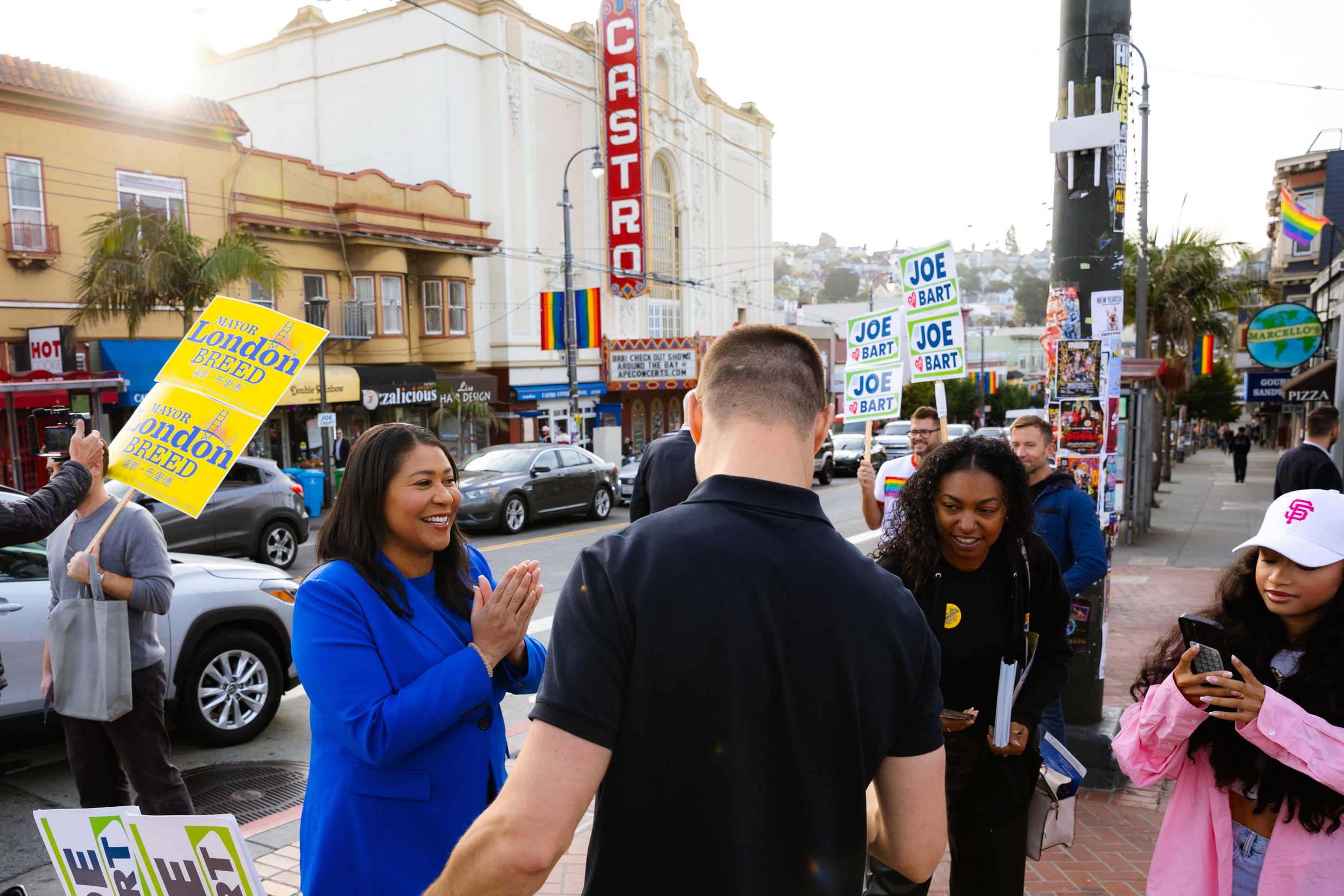 A group of people stand on a busy street, some holding campaign signs. A woman in a blue outfit smiles and talks, while others hold phones and observe.