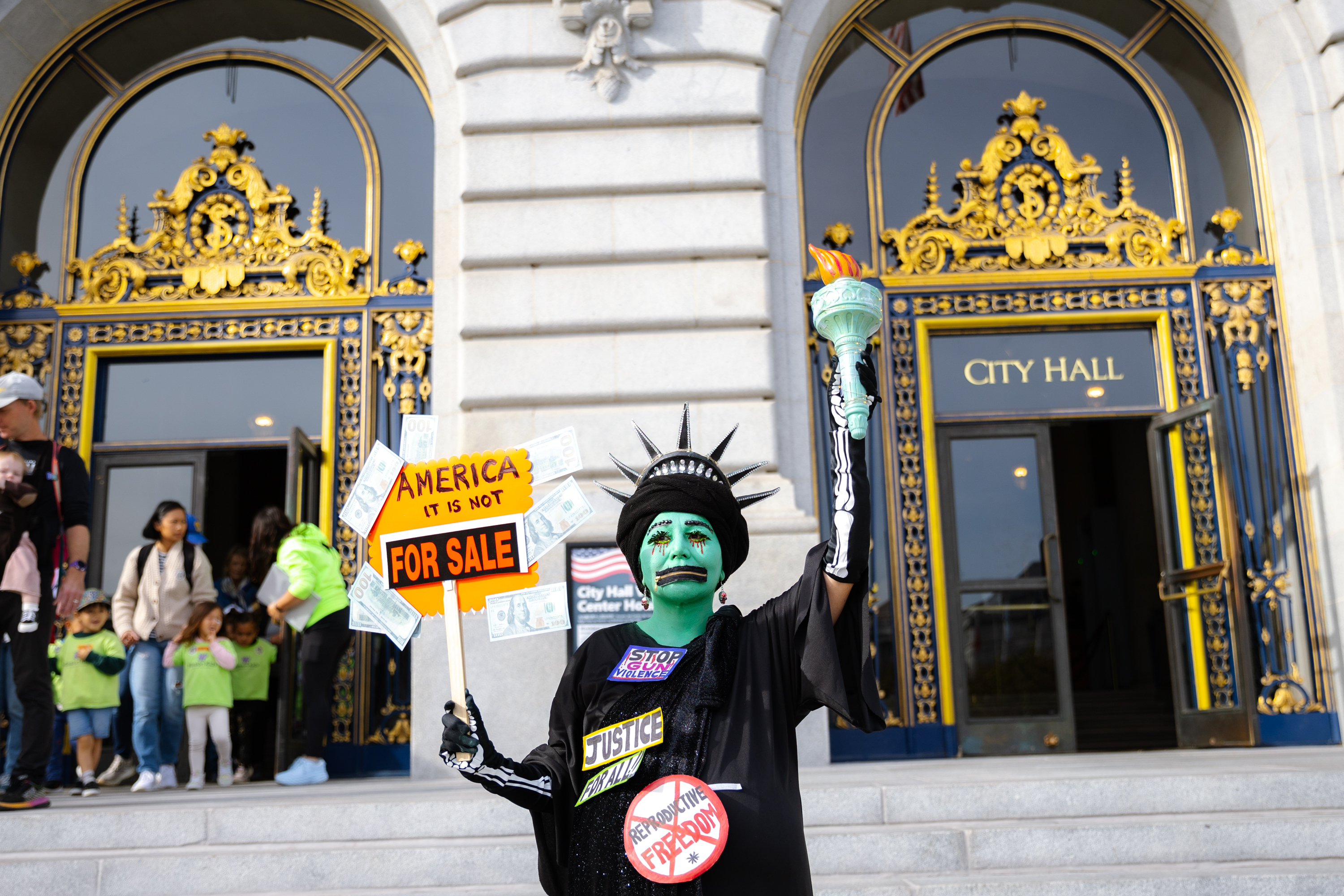 A person dressed as the Statue of Liberty holds a torch and a sign reading "America It Is Not For Sale" on the steps of a grand city hall entrance.