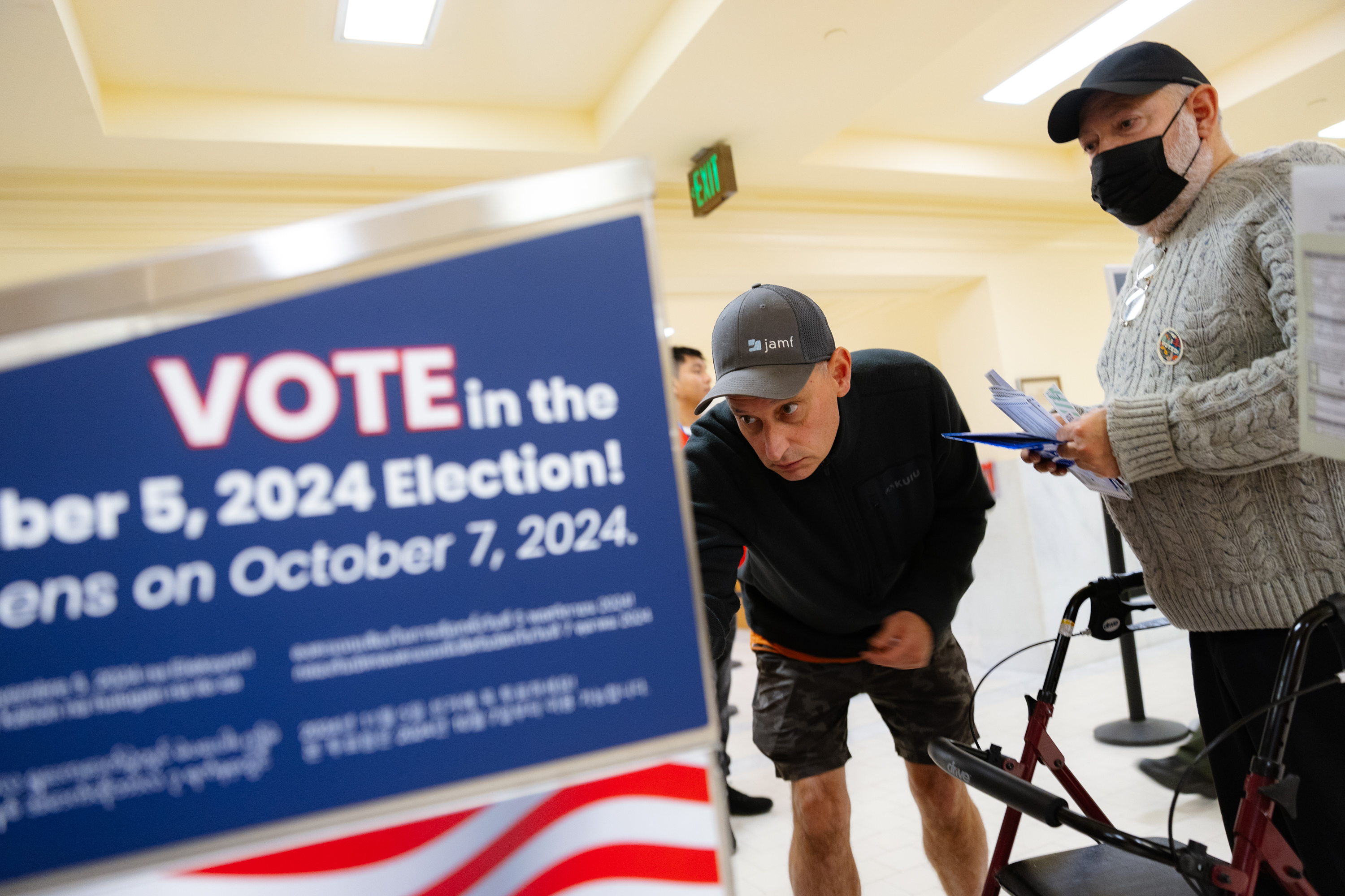 Two men are at a voting location. One man, wearing a cap and shorts, leans in to read a sign about the 2024 election. The other, in a mask, holds papers.