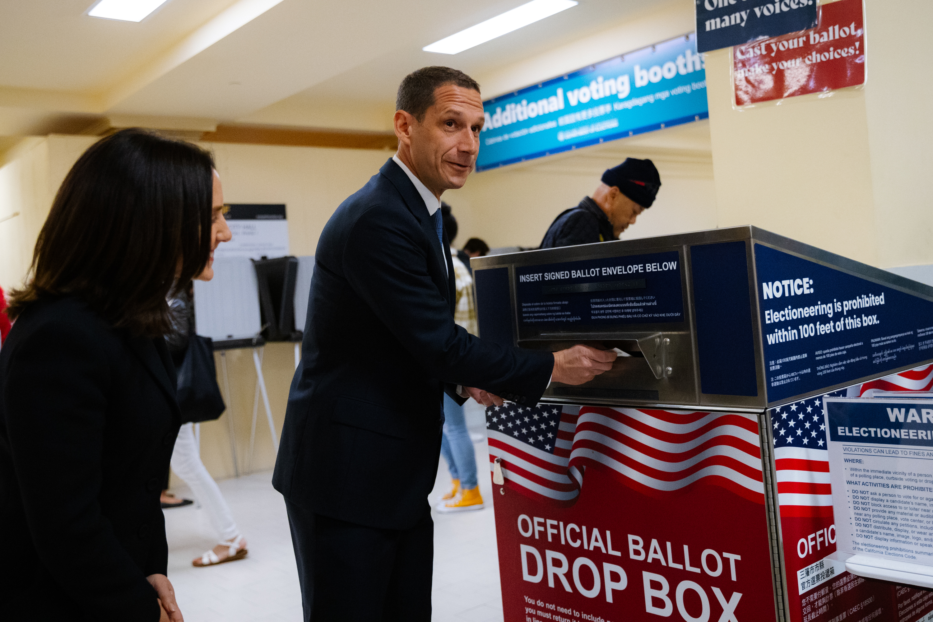 A man in a suit places an envelope in an official ballot drop box. A woman stands nearby, and there are signs on the wall about voting and election rules.