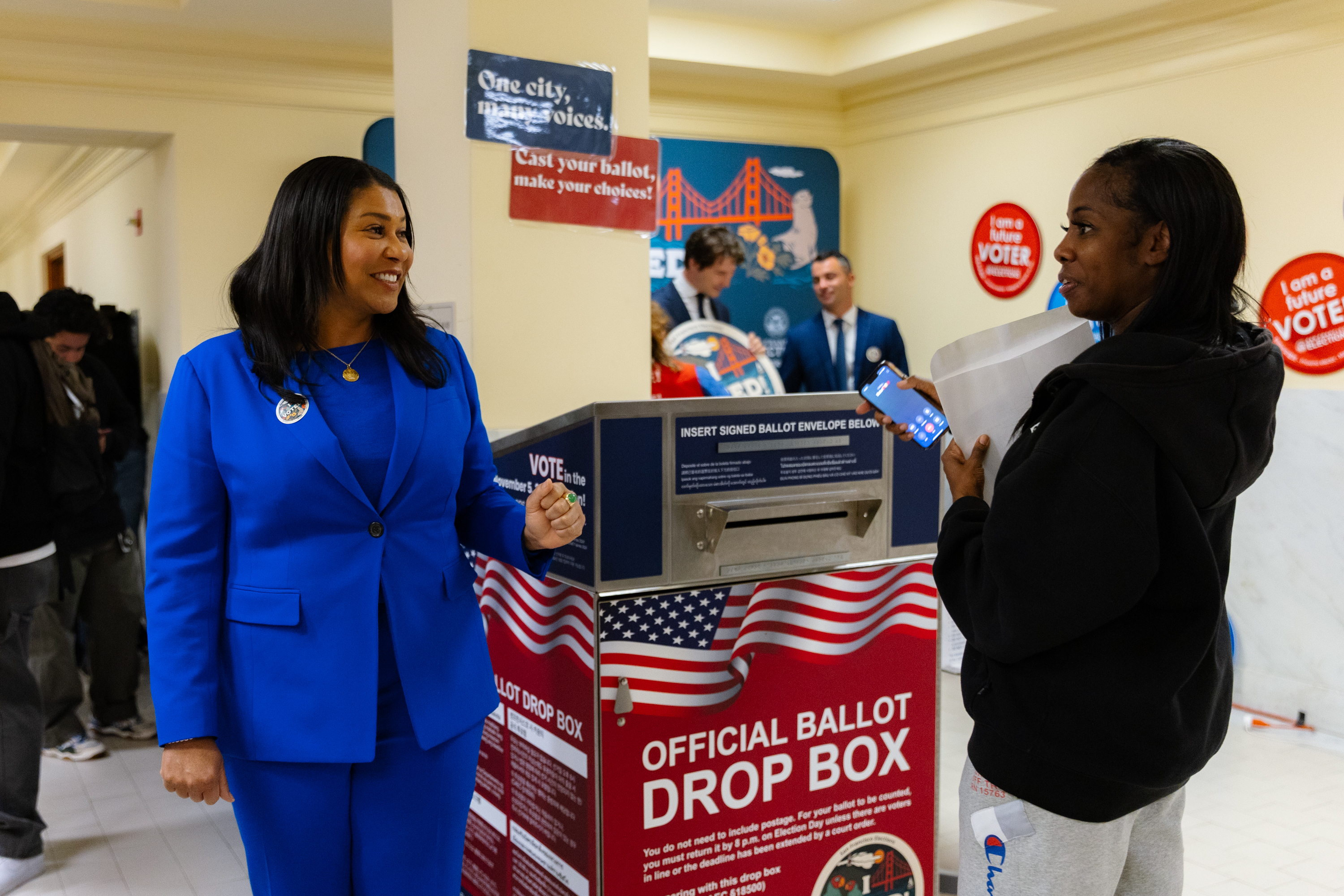 Two people are standing near an official ballot drop box. One is wearing a blue suit, smiling, and the other holds a phone and paper. Posters and people are in the background.