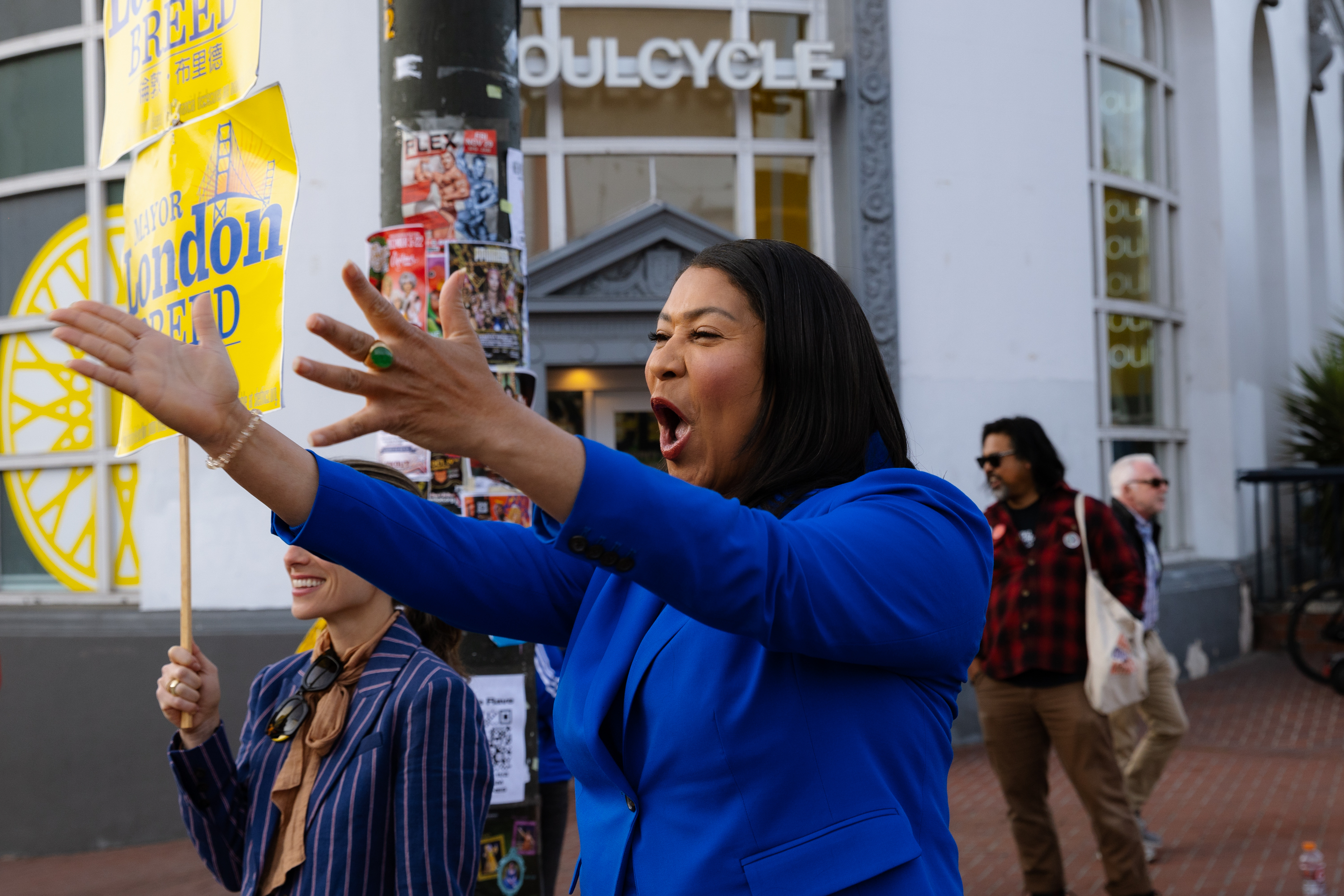 A woman in a blue suit enthusiastically gestures with open arms on a city street. She is in front of a building with "SoulCycle" signage. Others hold signs nearby.