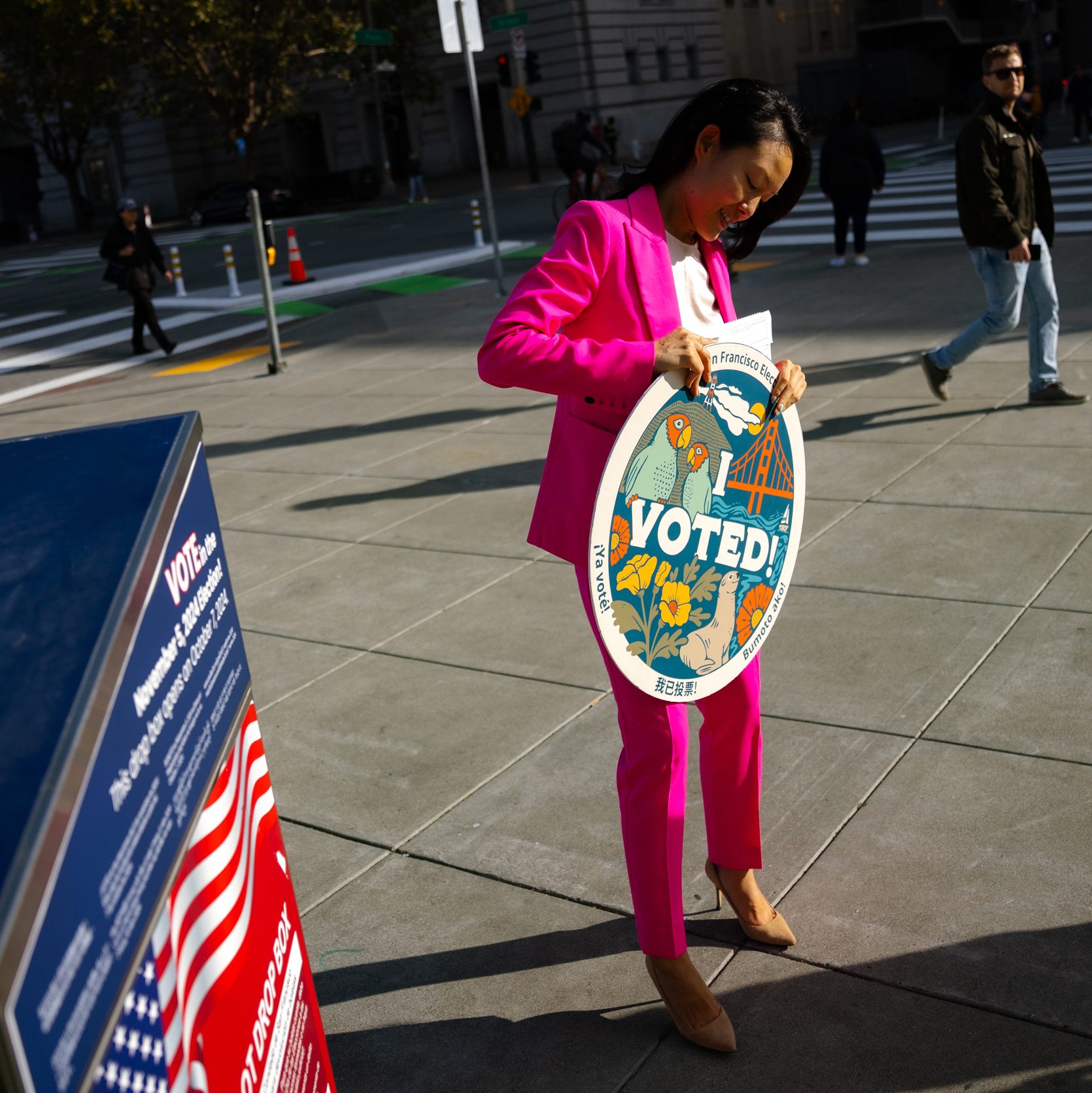 A woman in a bright pink suit stands on a city sidewalk, smiling at a colorful &quot;I Voted!&quot; sign. A ballot drop box with an American flag is nearby.