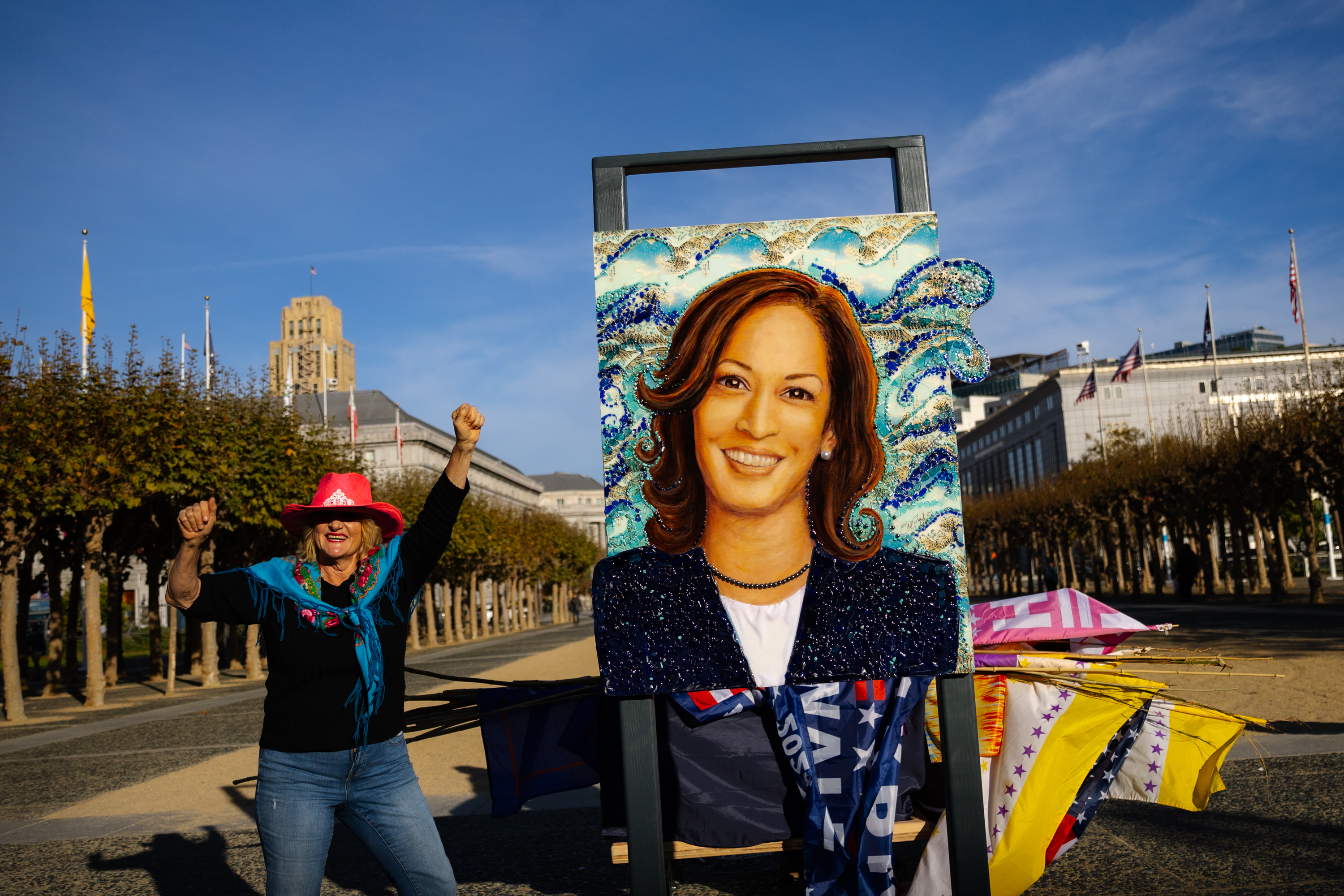 A person in a red hat and blue scarf raises their arms joyfully beside a vibrant portrait on an easel, with building and flags in the background.