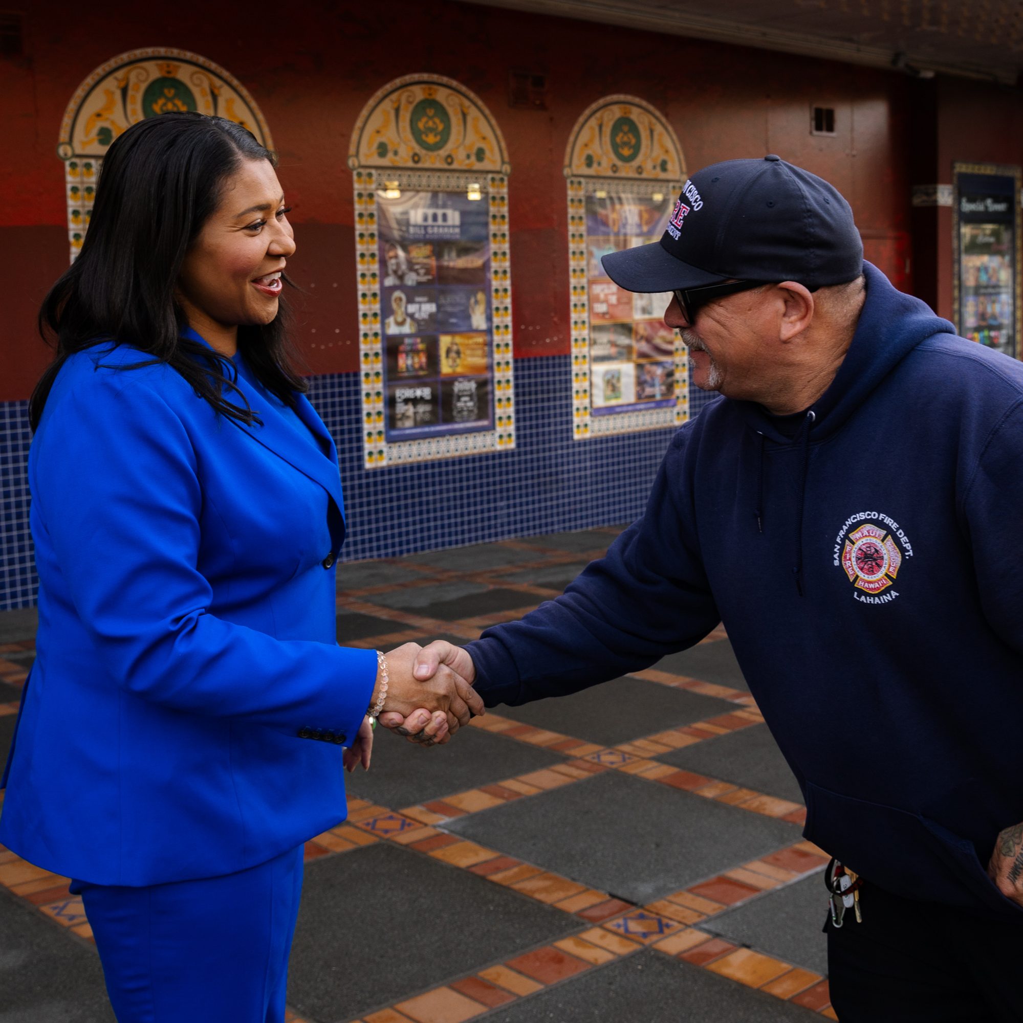 A woman in a bright blue suit is shaking hands with a man in a navy blue hoodie and cap. They are standing outside a building with decorative tiles.