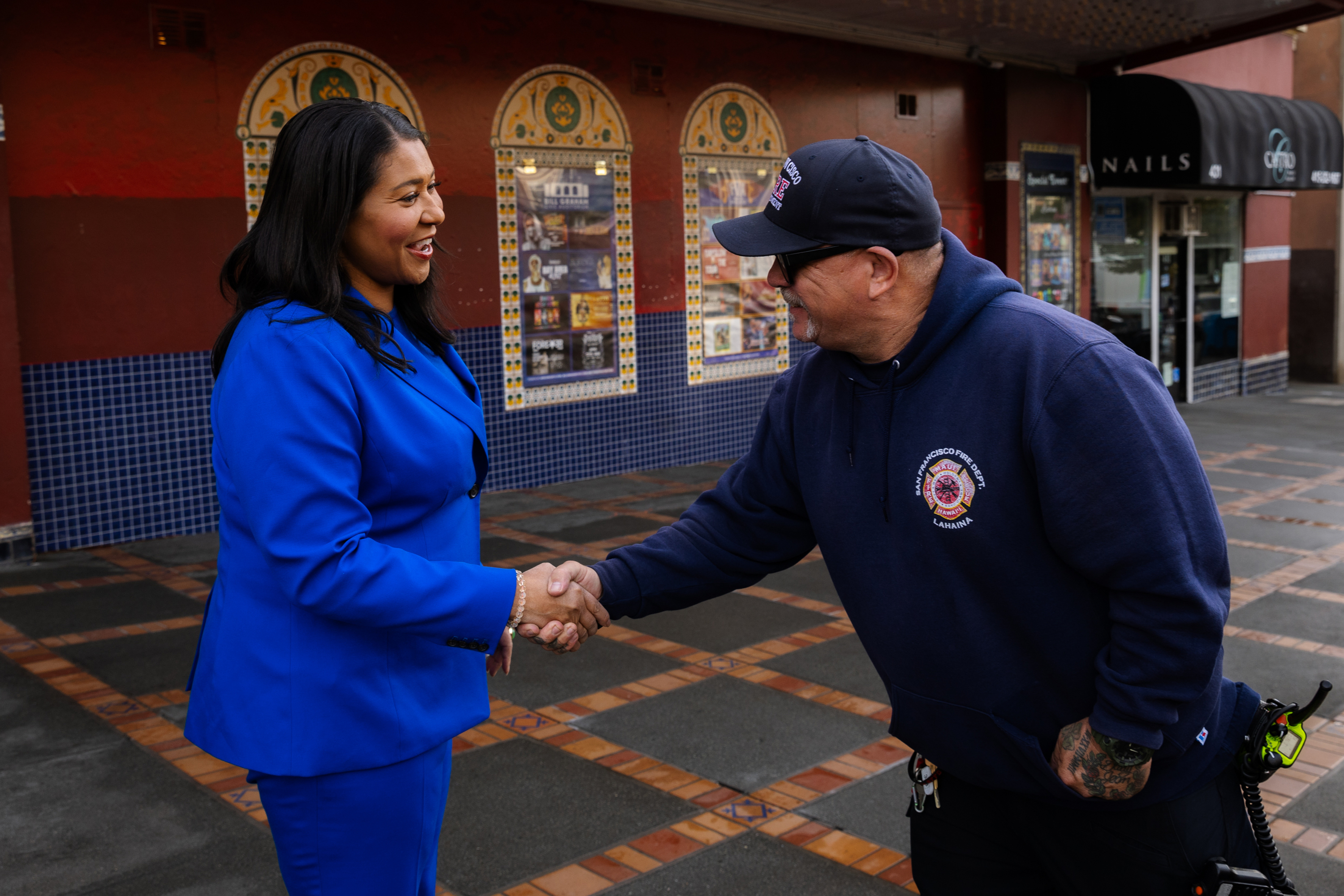 A woman in a bright blue suit is shaking hands with a man in a navy blue hoodie and cap. They are standing outside a building with decorative tiles.