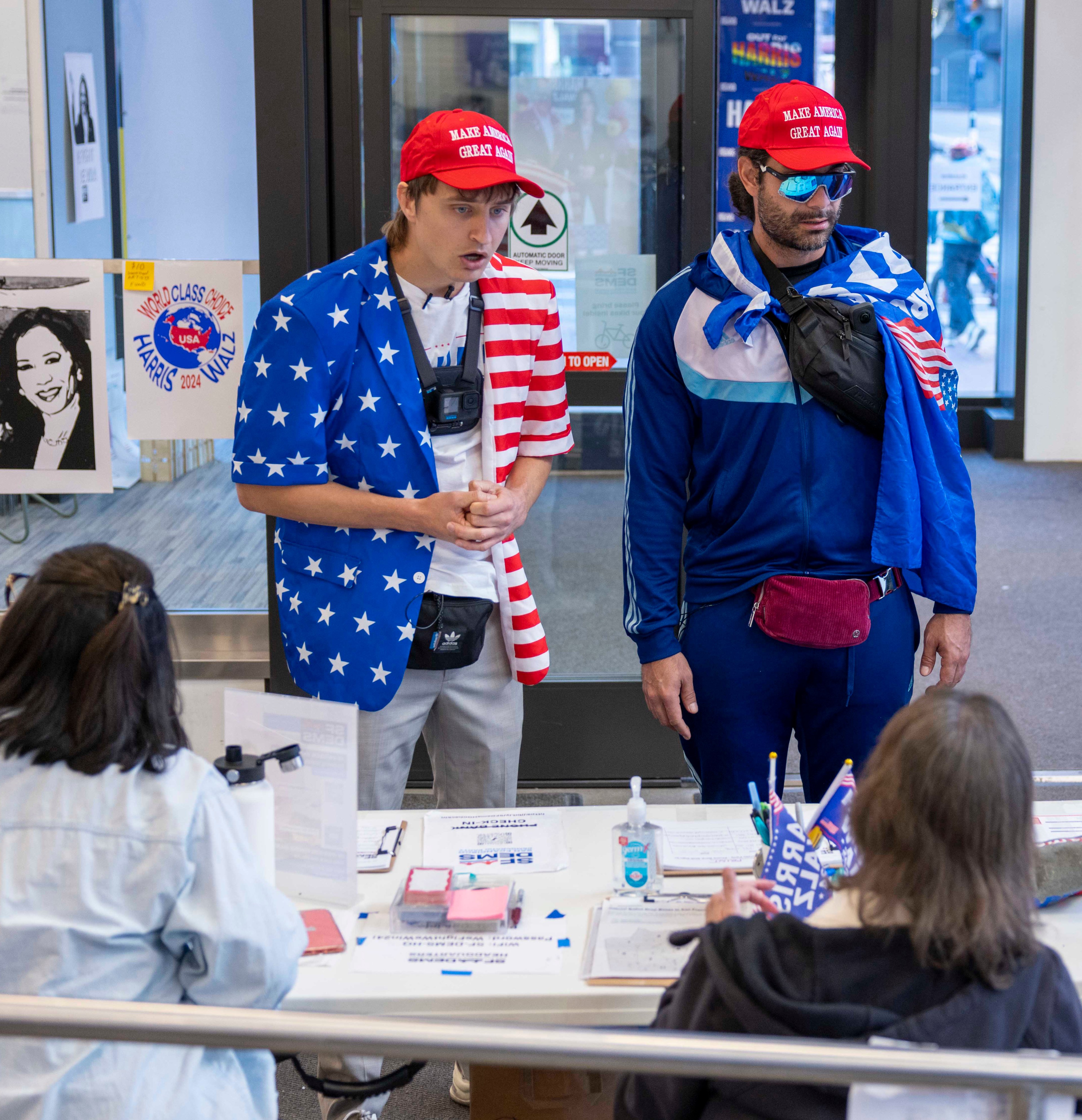 Two people in patriotic attire and red hats stand at a table with documents and voting paraphernalia. A portrait of a woman is on display.