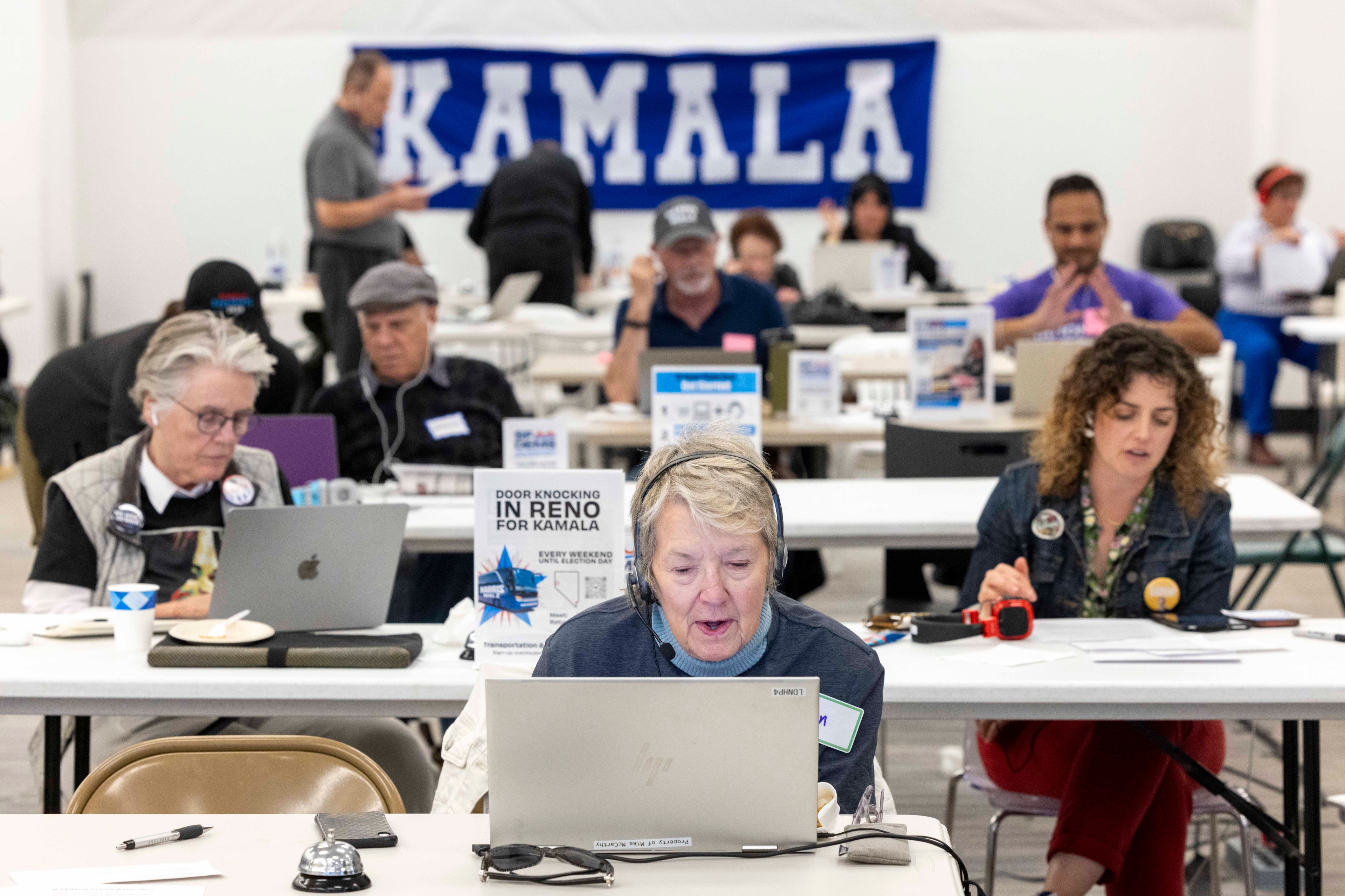 A group of people are working at tables with laptops in a room. A large sign with &quot;Kamala&quot; is visible in the background, and there's a poster about door knocking.