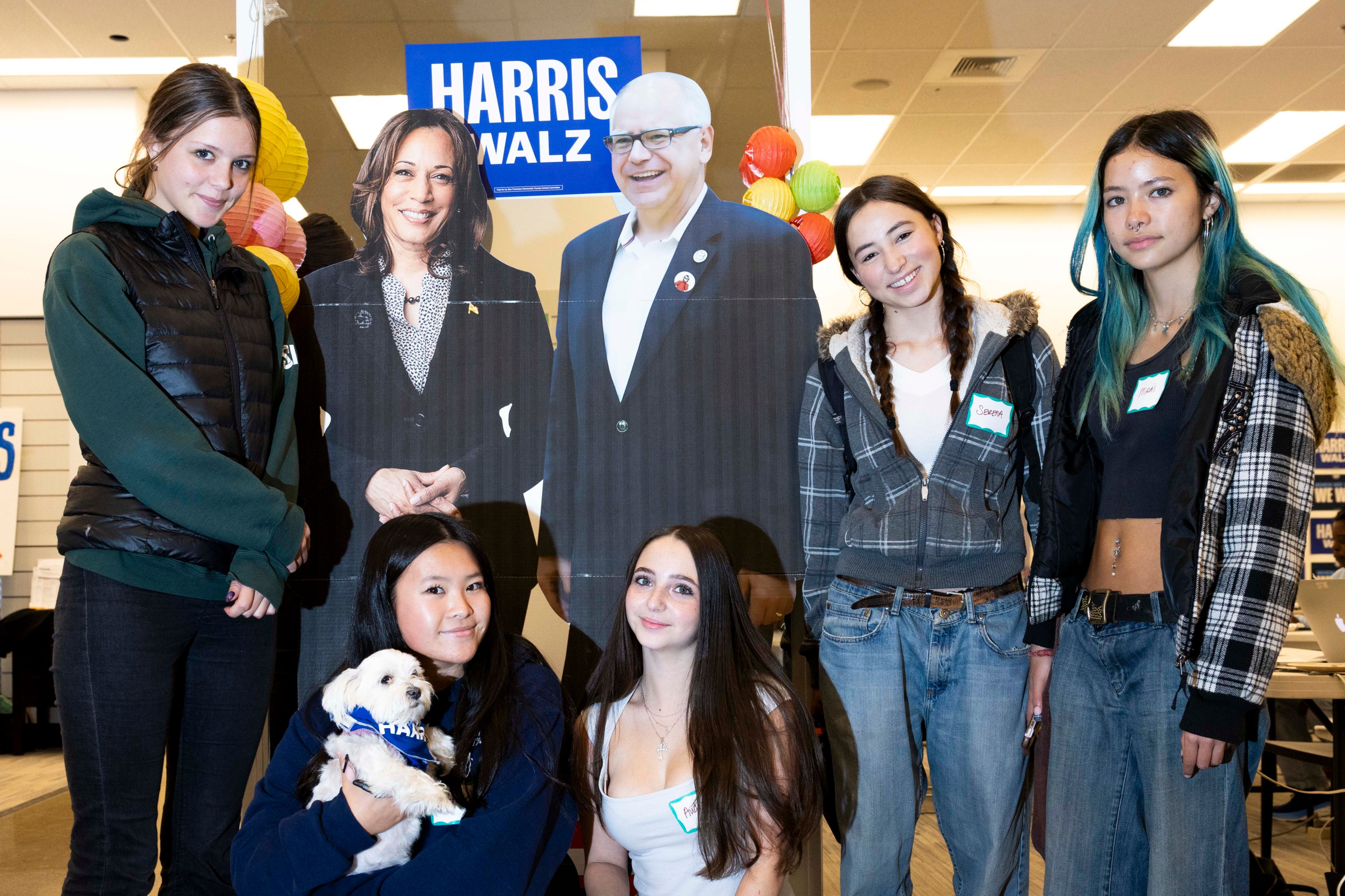 Five young women stand around cardboard cutouts of two political figures, smiling. One holds a small dog. Posters with &quot;Harris Walz&quot; are in the background.