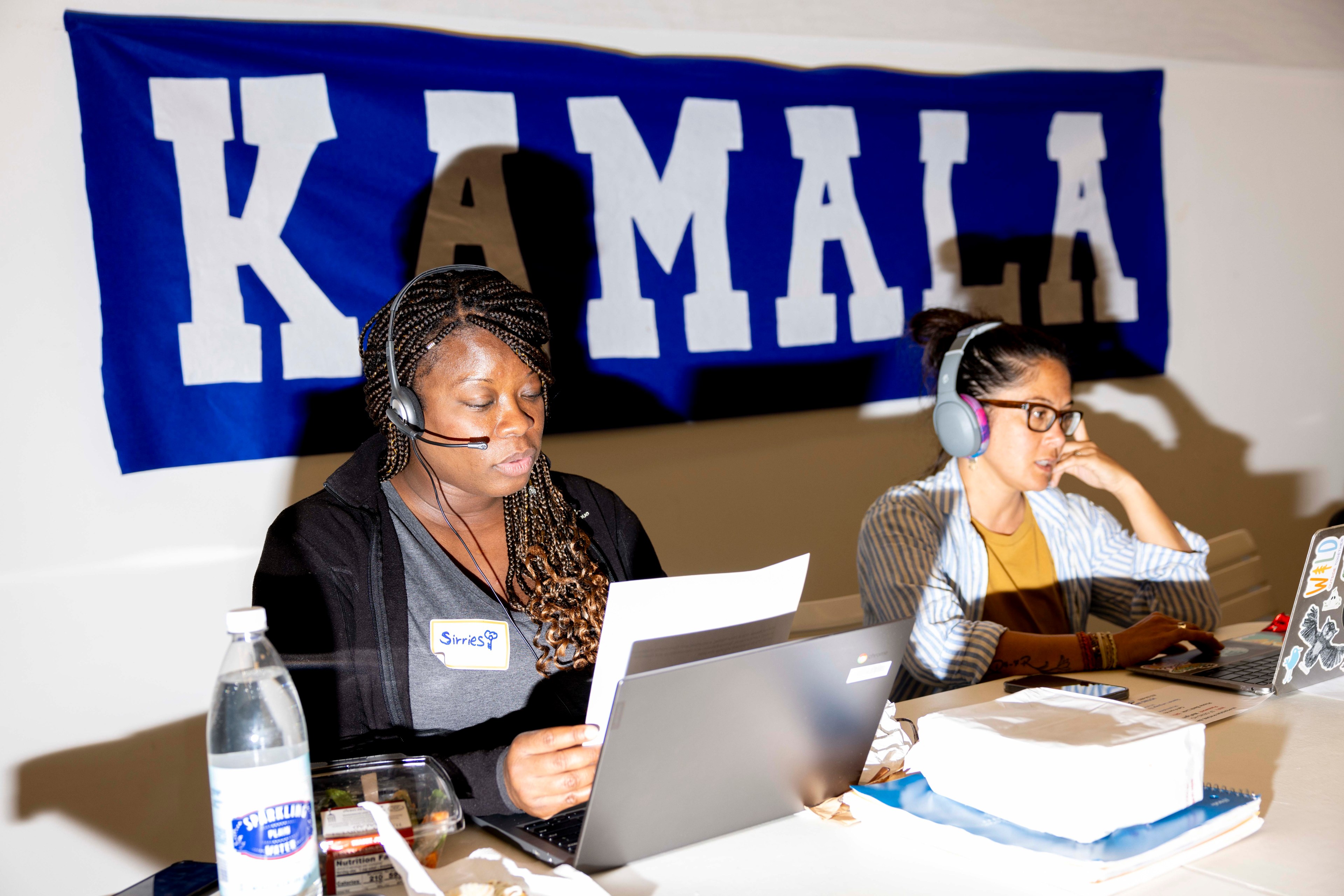 Two people sit at a table with laptops and headsets in front of a &quot;KAMALA&quot; banner, engaged in phone work. A water bottle and papers are on the table.