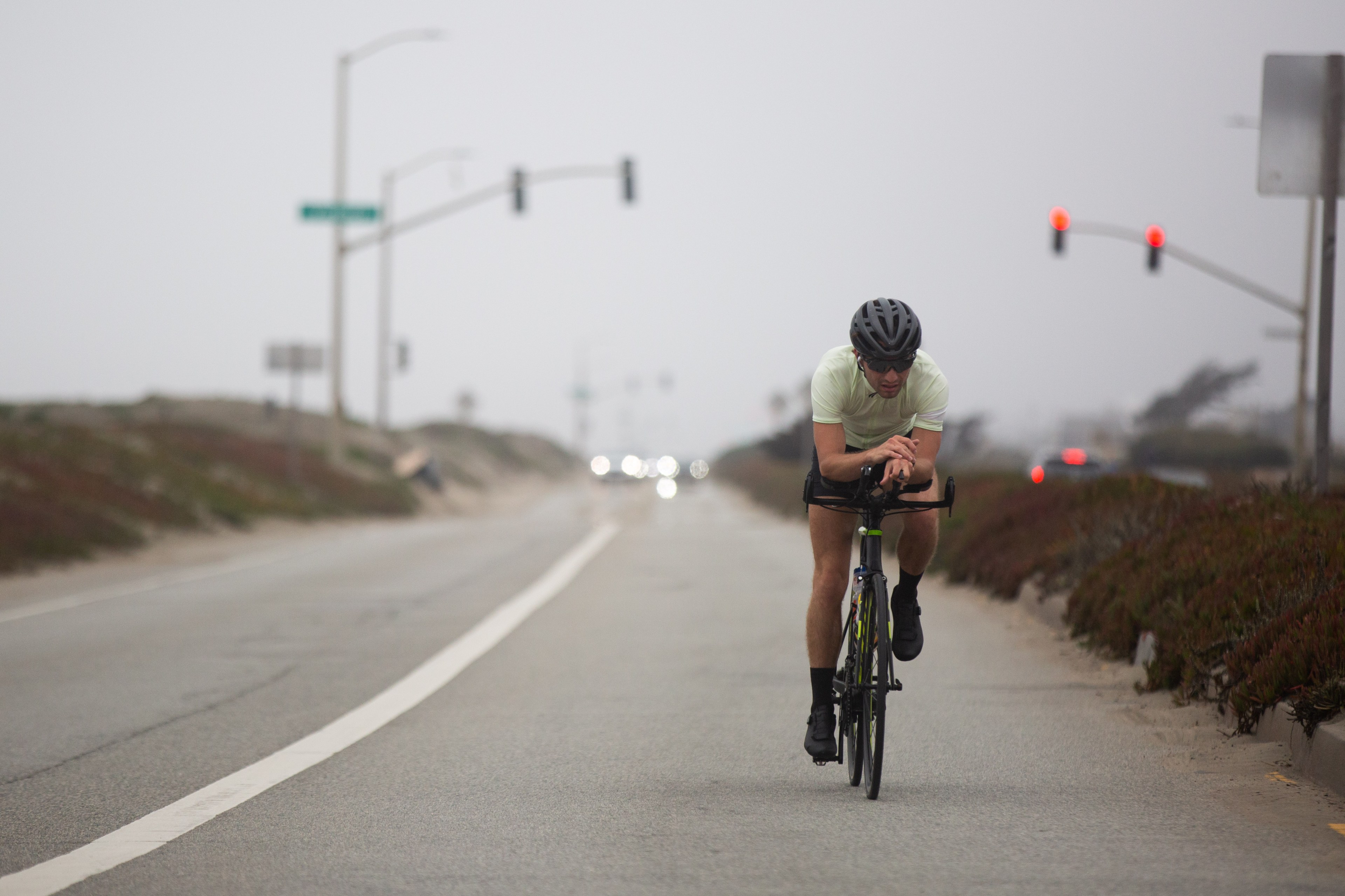 A cyclist wearing a helmet and light shirt rides on a deserted road under cloudy skies. Traffic lights in the distance add a touch of color to the scene.