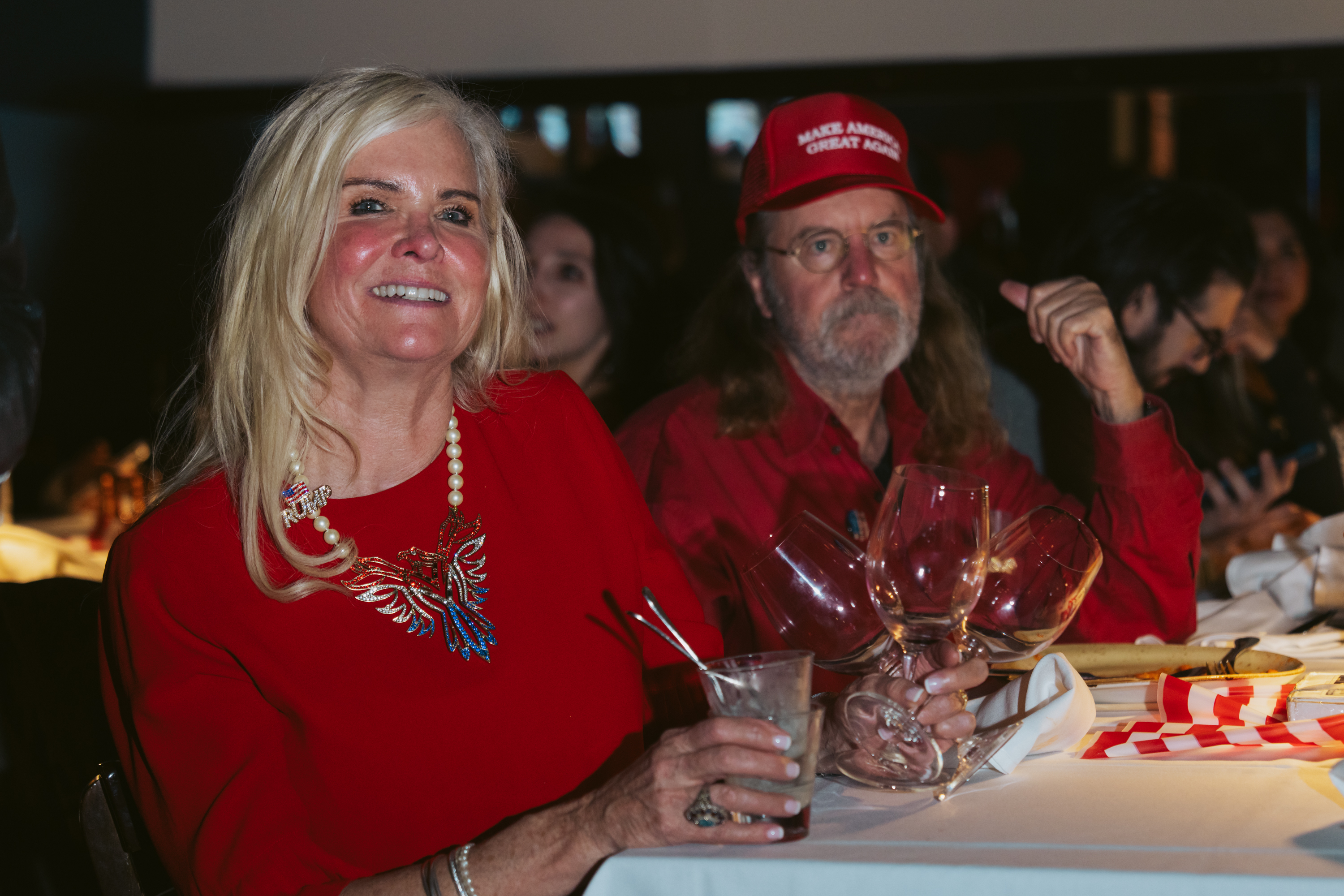 A smiling woman in a red outfit and necklace holds a drink. Beside her, a man with a red cap sits at a table with wine glasses and napkins.