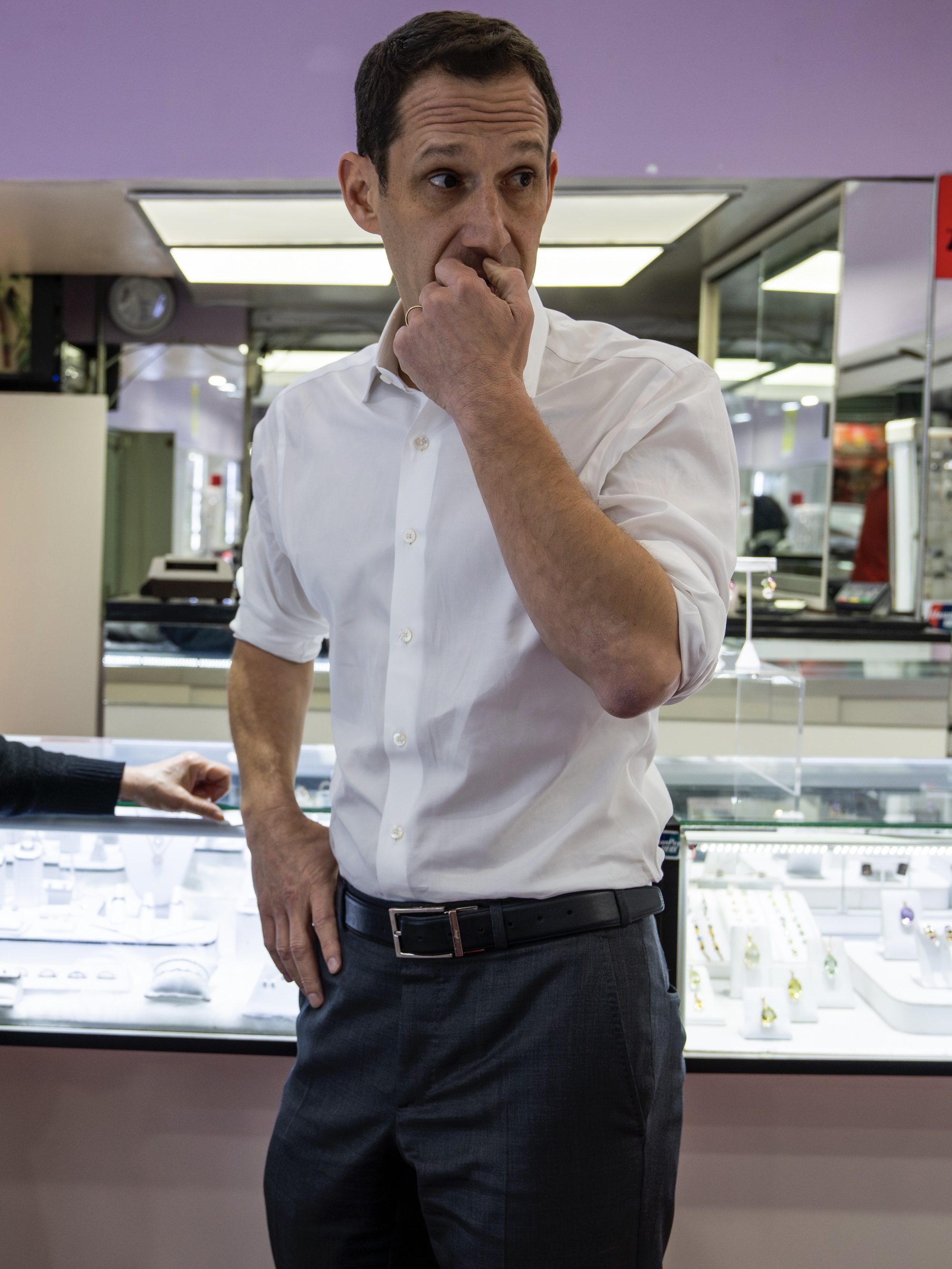 A man in a white shirt stands pensively in a jewelry store, with display cases of rings and necklaces behind him and a hand pointing at one case.