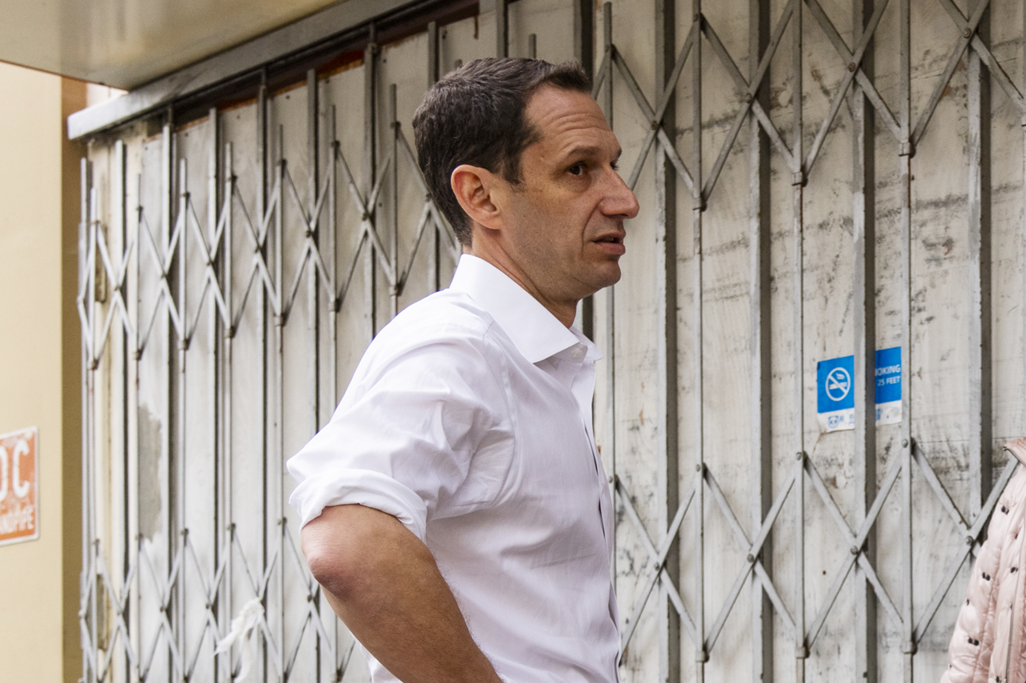 A man in a white shirt stands in front of a closed metal gate with his hands on his hips. A blue "no smoking" sign is visible in the background.